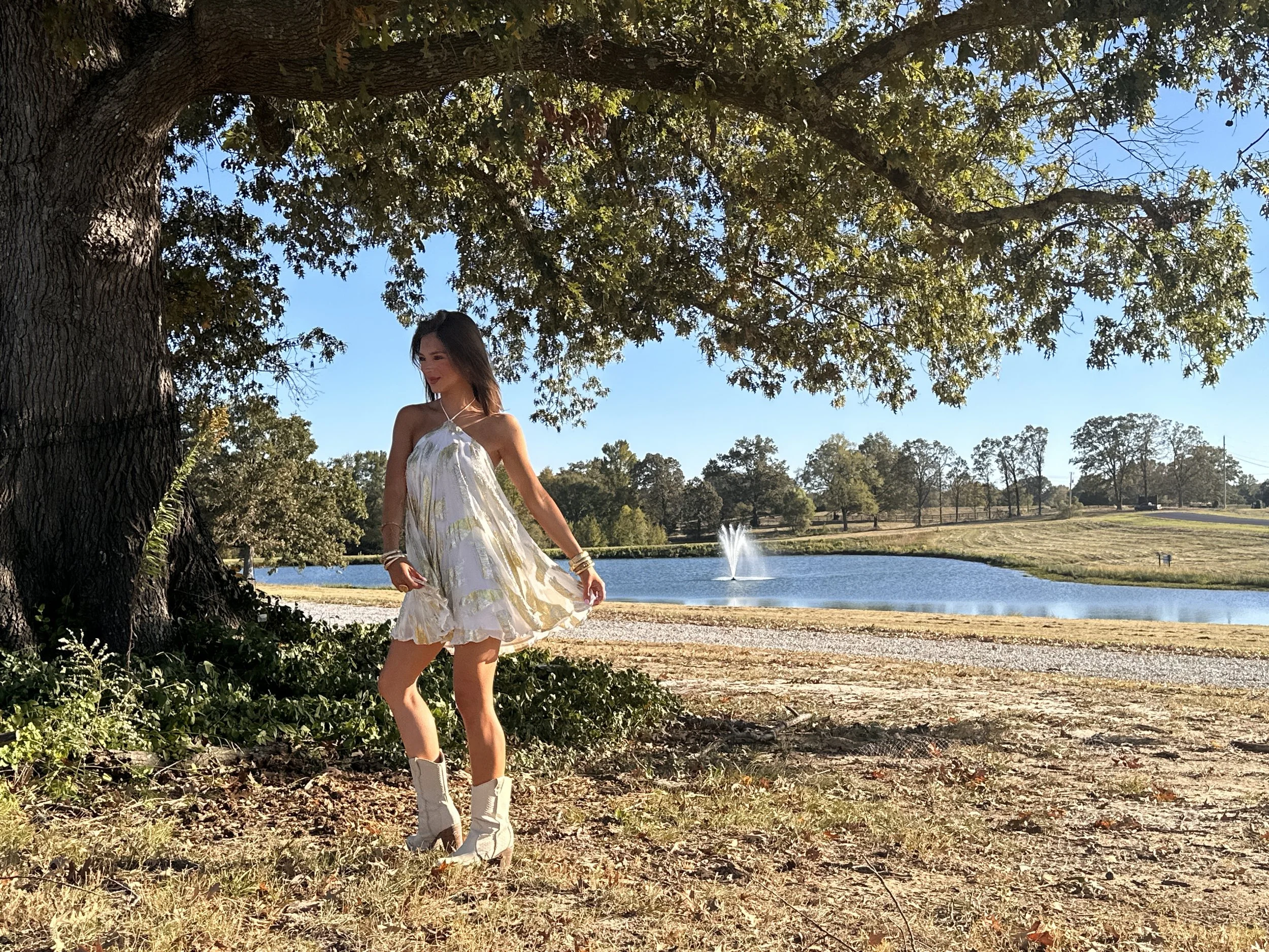 Young woman in a white dress and cowboy boots standing outdoors near a large tree, with a pond and fountain in the background on a sunny day.