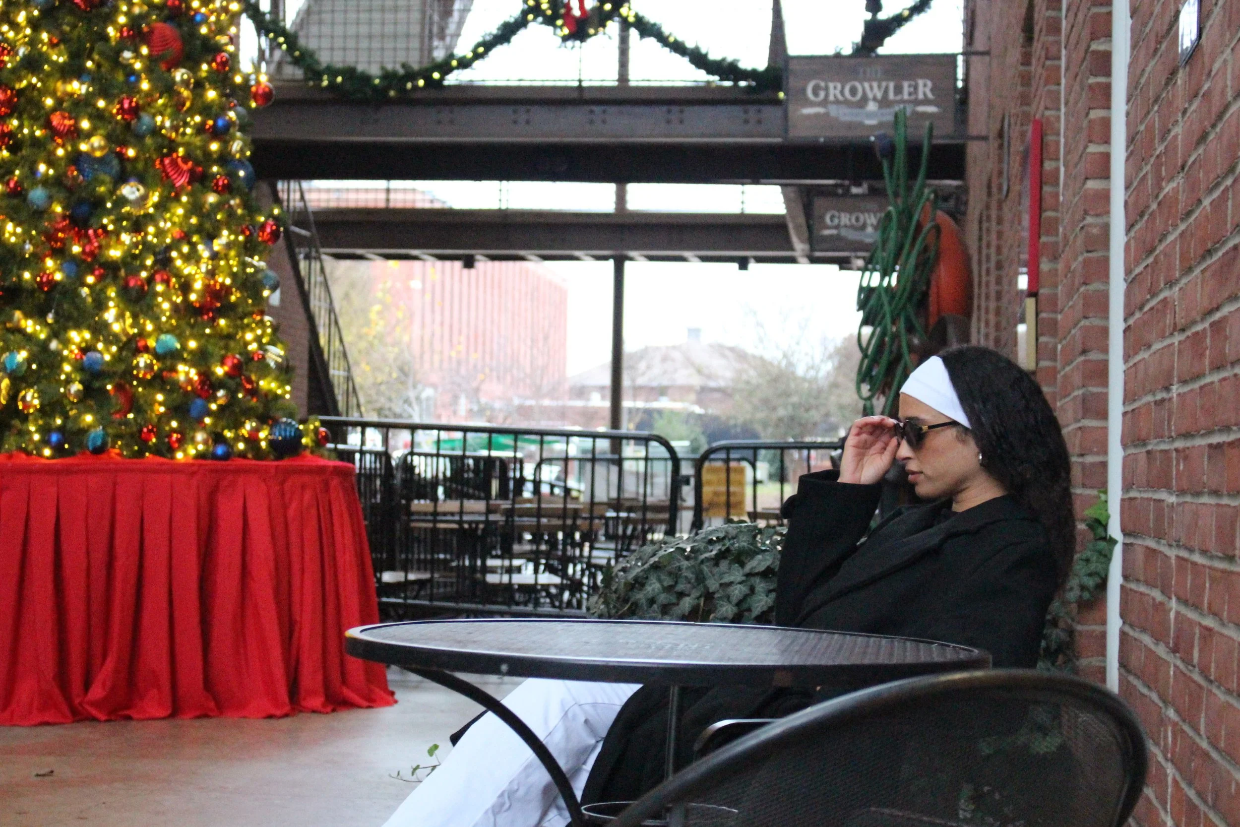 A woman with glasses and a white headband sitting beside a Christmas tree decorated with lights and ornaments, near a brick wall and outdoor area.