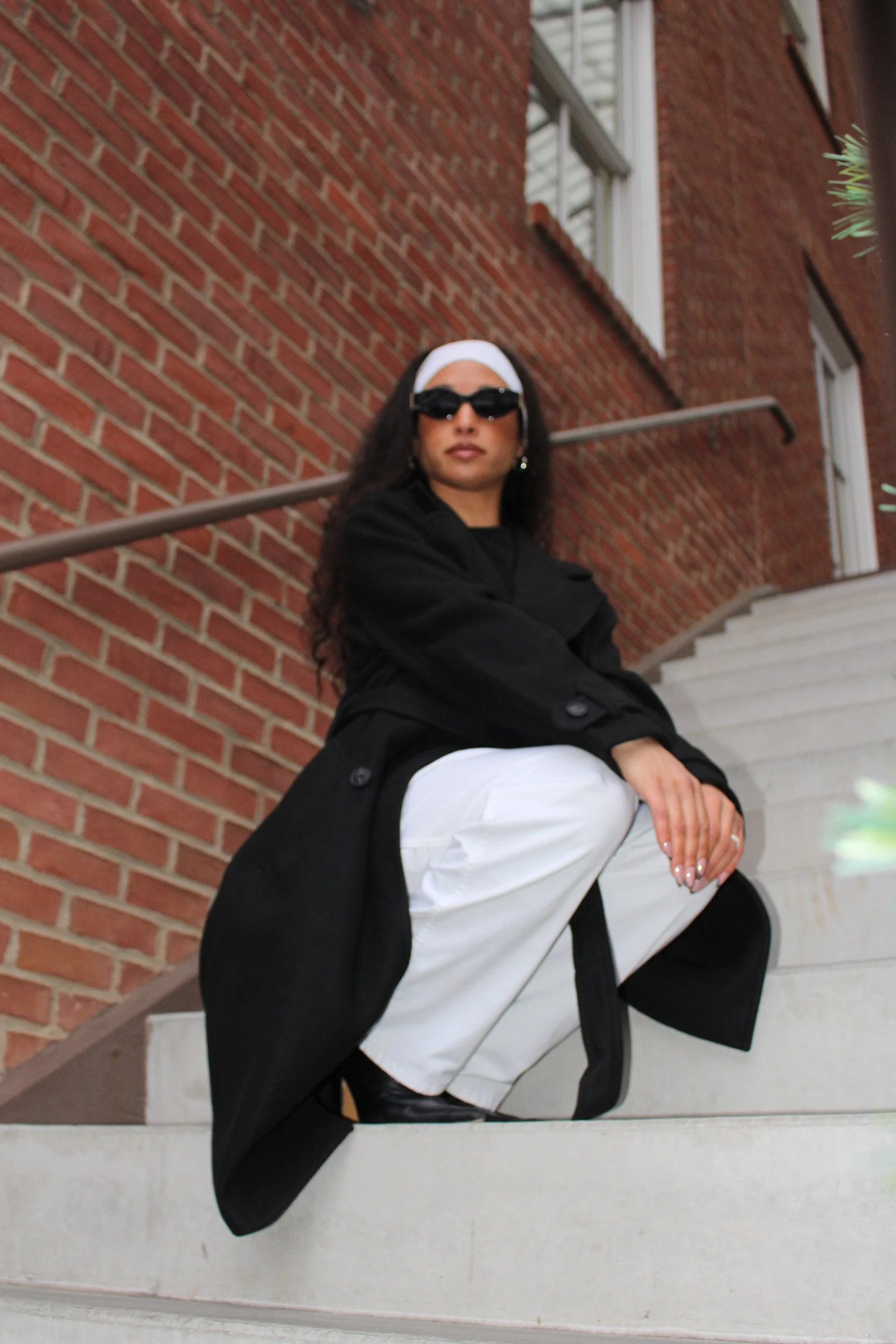 Person with long curly hair, wearing sunglasses, a white headband, black coat, and white pants, sitting on white stairs in front of a brick wall with windows.