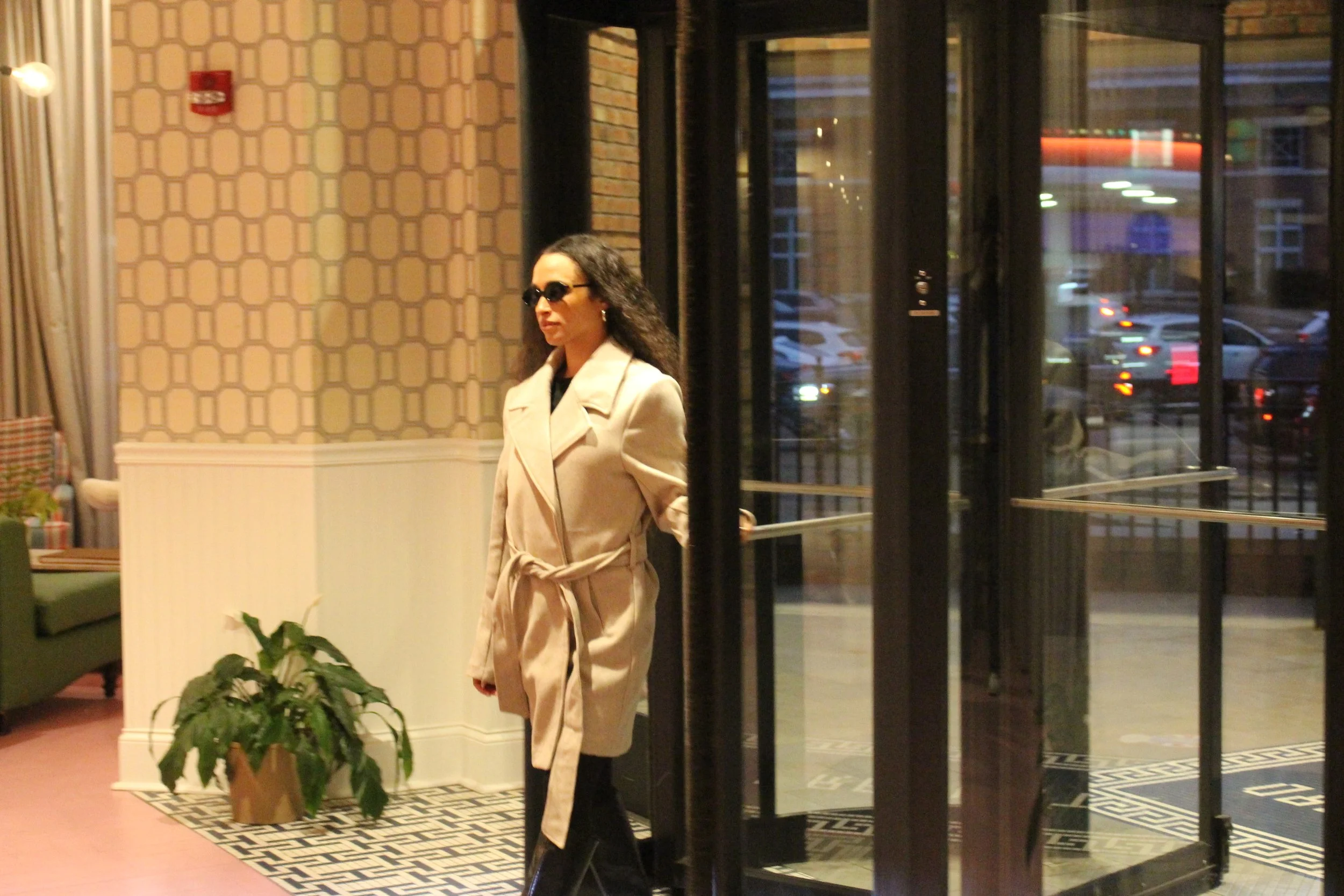 Woman with curly hair wearing a beige trench coat and sunglasses standing inside a building near glass doors, with city lights visible outside.