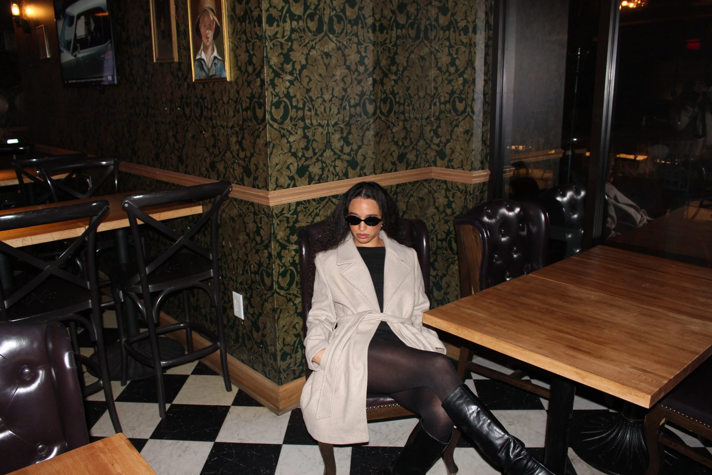 A woman with curly hair wearing sunglasses, a beige coat, black pants, and black boots sitting in a restaurant or cafe with wooden tables and dark chairs, and a patterned wallpaper background.