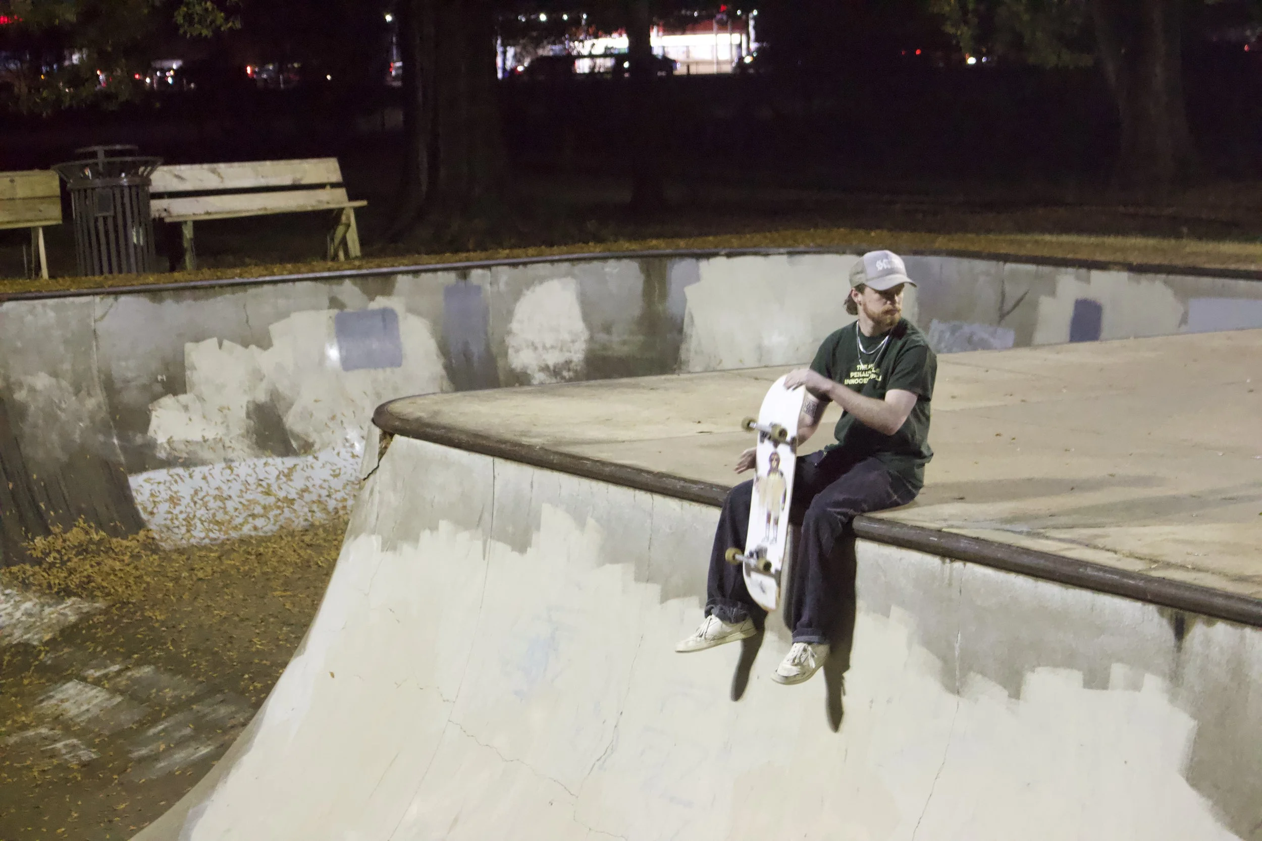 A young man with a beard and sunglasses sitting on the edge of a skatepark bowl at night. He is holding a skateboard and wearing a cap, black T-shirt, and dark pants. There are benches and a trash can in the background.
