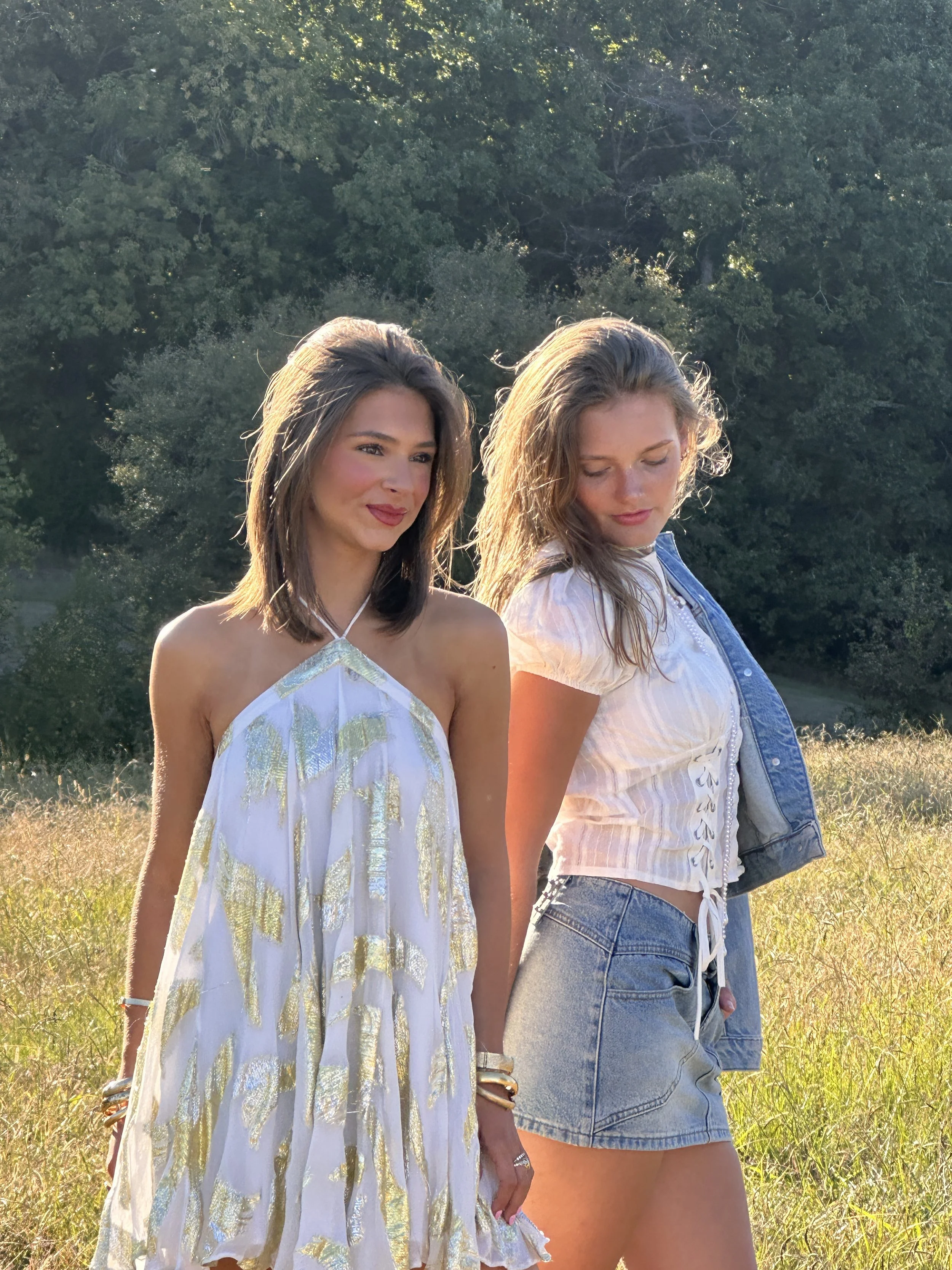 Two women standing outdoors in a grassy field, with trees in the background and sunlight shining on them, one with dark hair in a white dress and the other with light hair in a denim jacket and shorts.