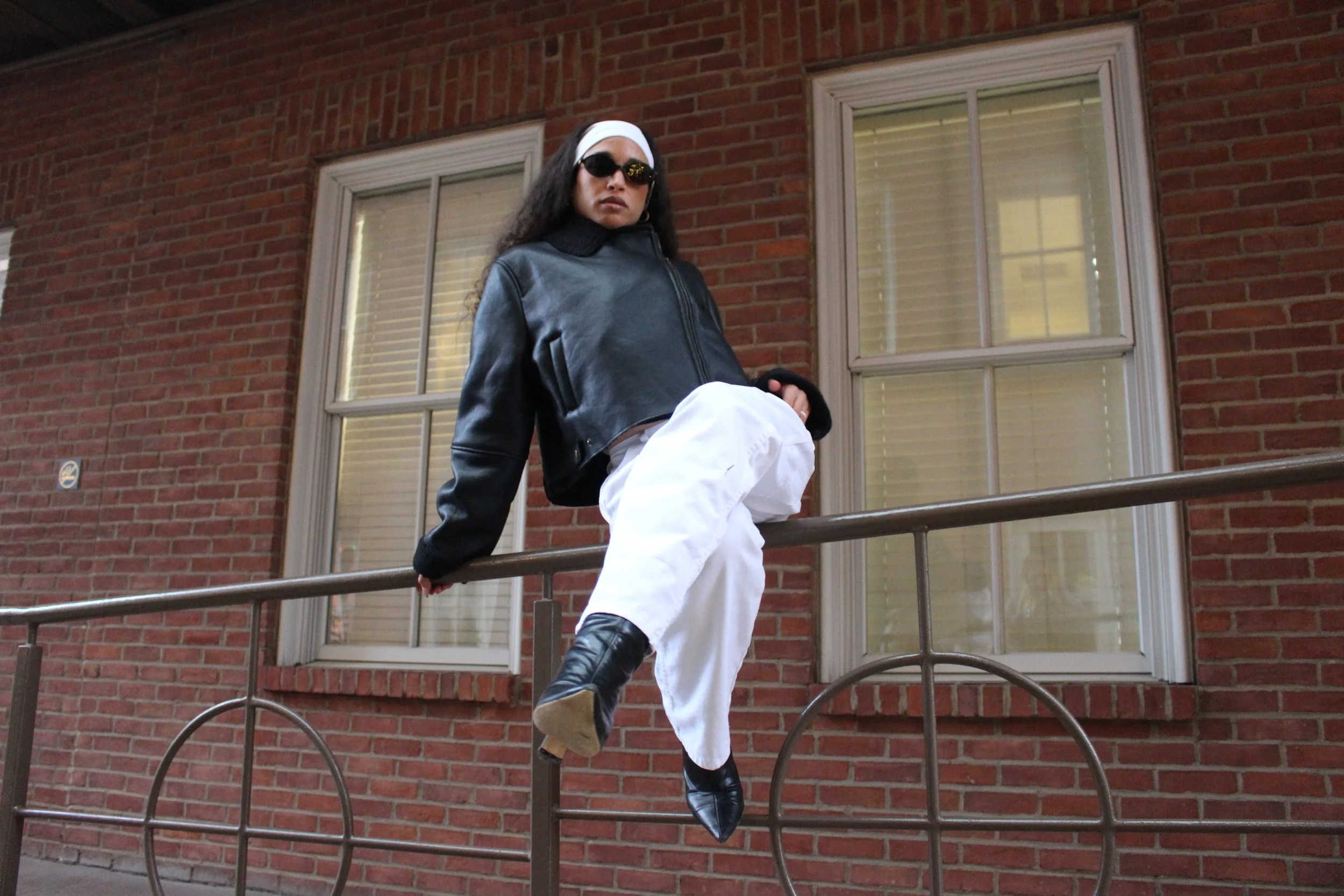 A woman with long dark hair, wearing sunglasses, a white headband, a black leather jacket, white pants, and black boots, standing on a metal railing in front of a red brick building with windows.