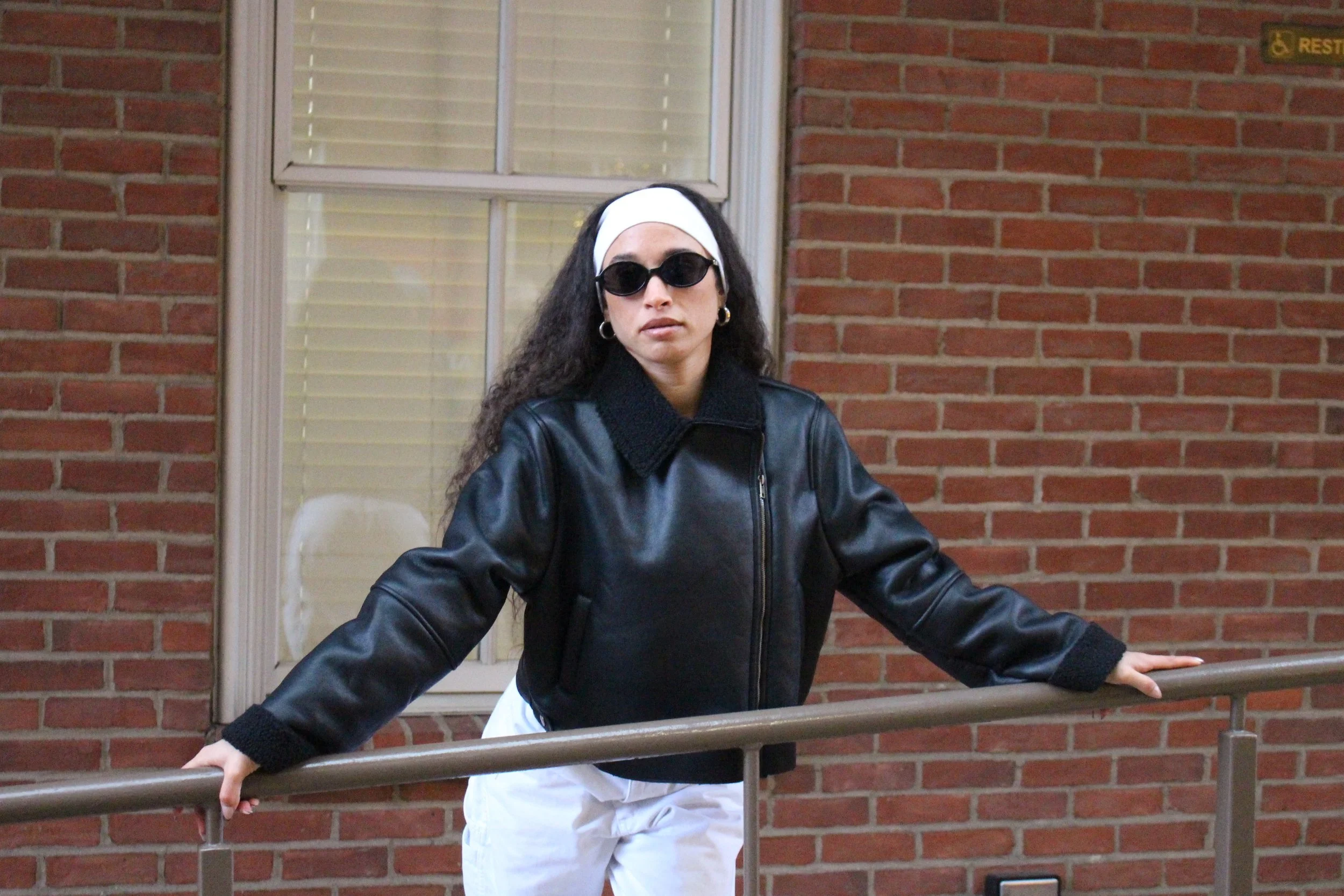 A woman with dark curly hair, wearing black sunglasses, white headband, gold hoop earrings, a black leather jacket, white pants, and black gloves, stands with arms outstretched on a metal railing in front of a brick building with a large window.