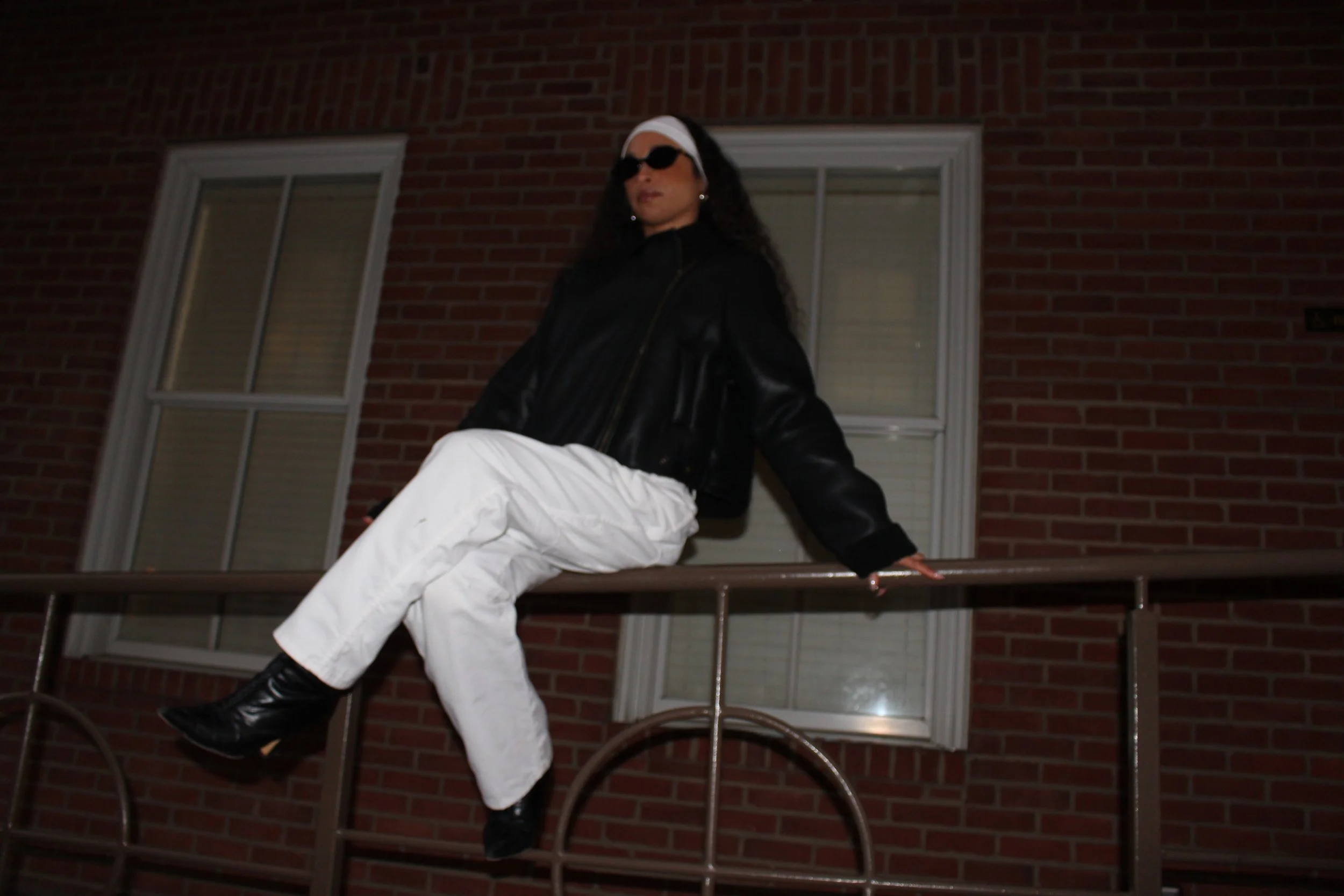 A woman sitting on a balcony railing at night, dressed in white pants, a black leather jacket, black boots, dark sunglasses, a white headscarf, and earrings, against a brick building with two windows.