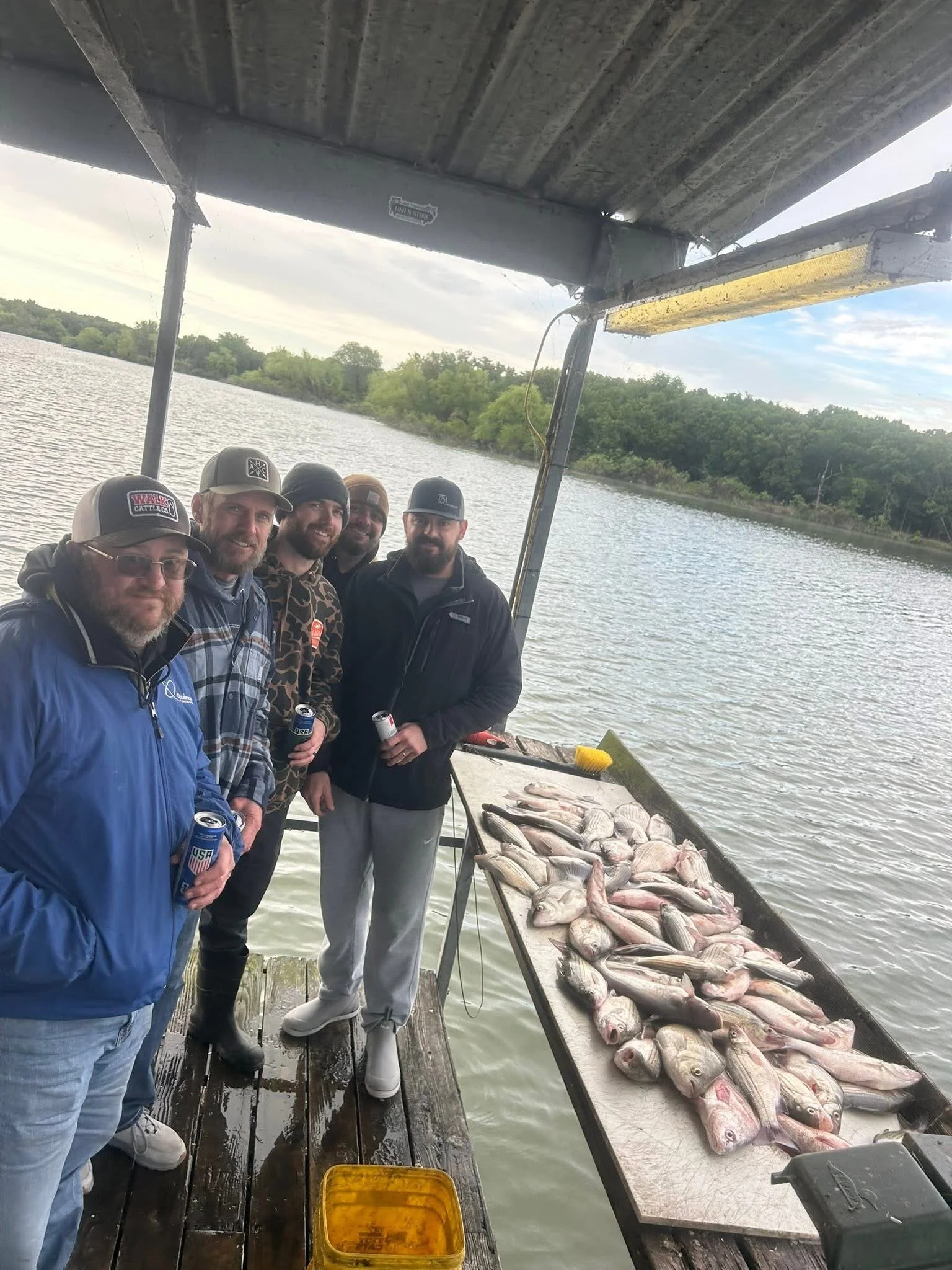 Five men are on a boat by a river, displaying their catch of fish on a table. They are dressed in casual clothing, some with hats and jackets, holding cans of drinks, and smiling at the camera. The tree-lined river bank is visible in the background, 