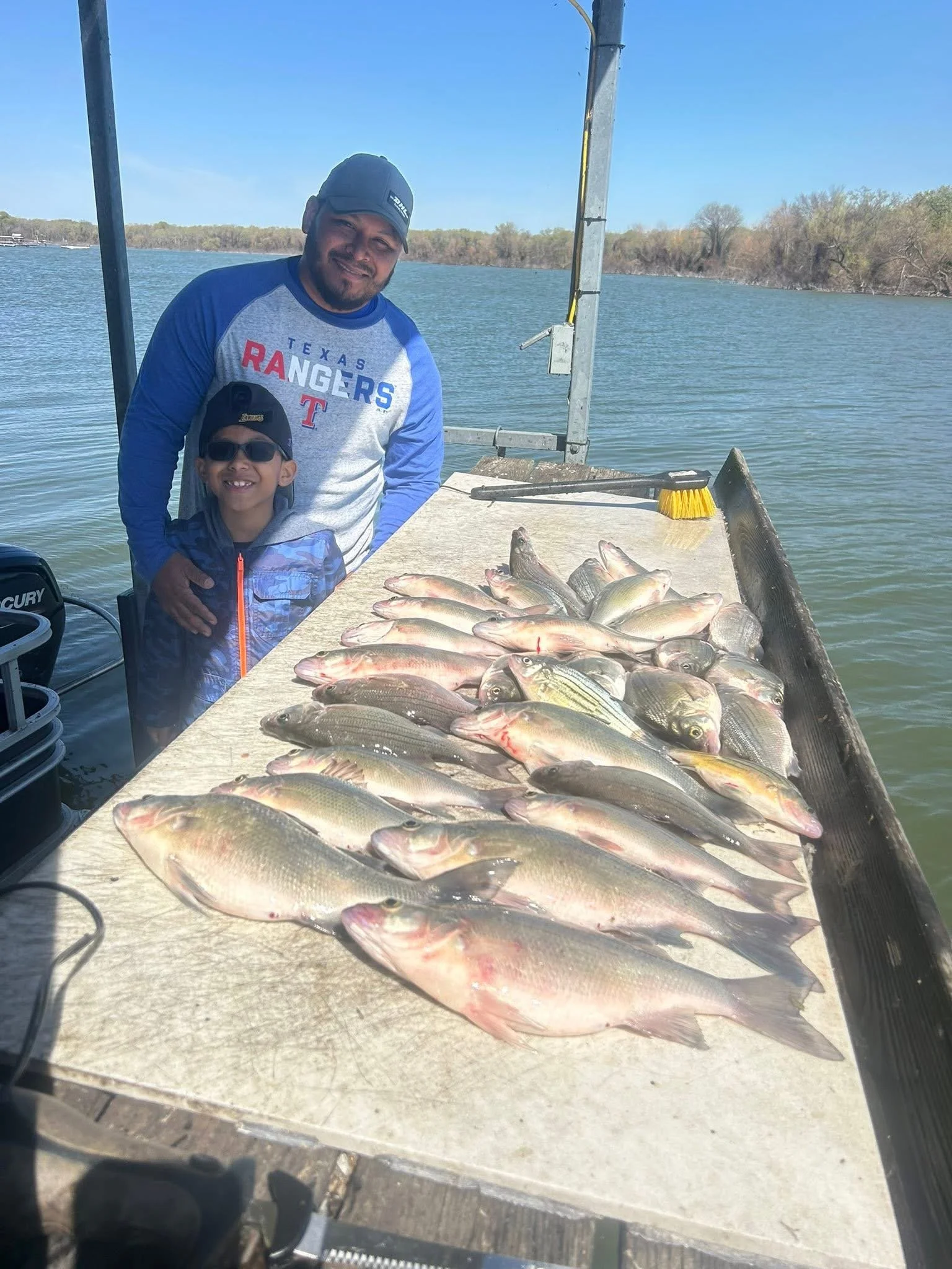 Two smiling people, an adult man and a young boy, stand behind a table filled with freshly caught fish on a boat by a body of water with trees in the background.