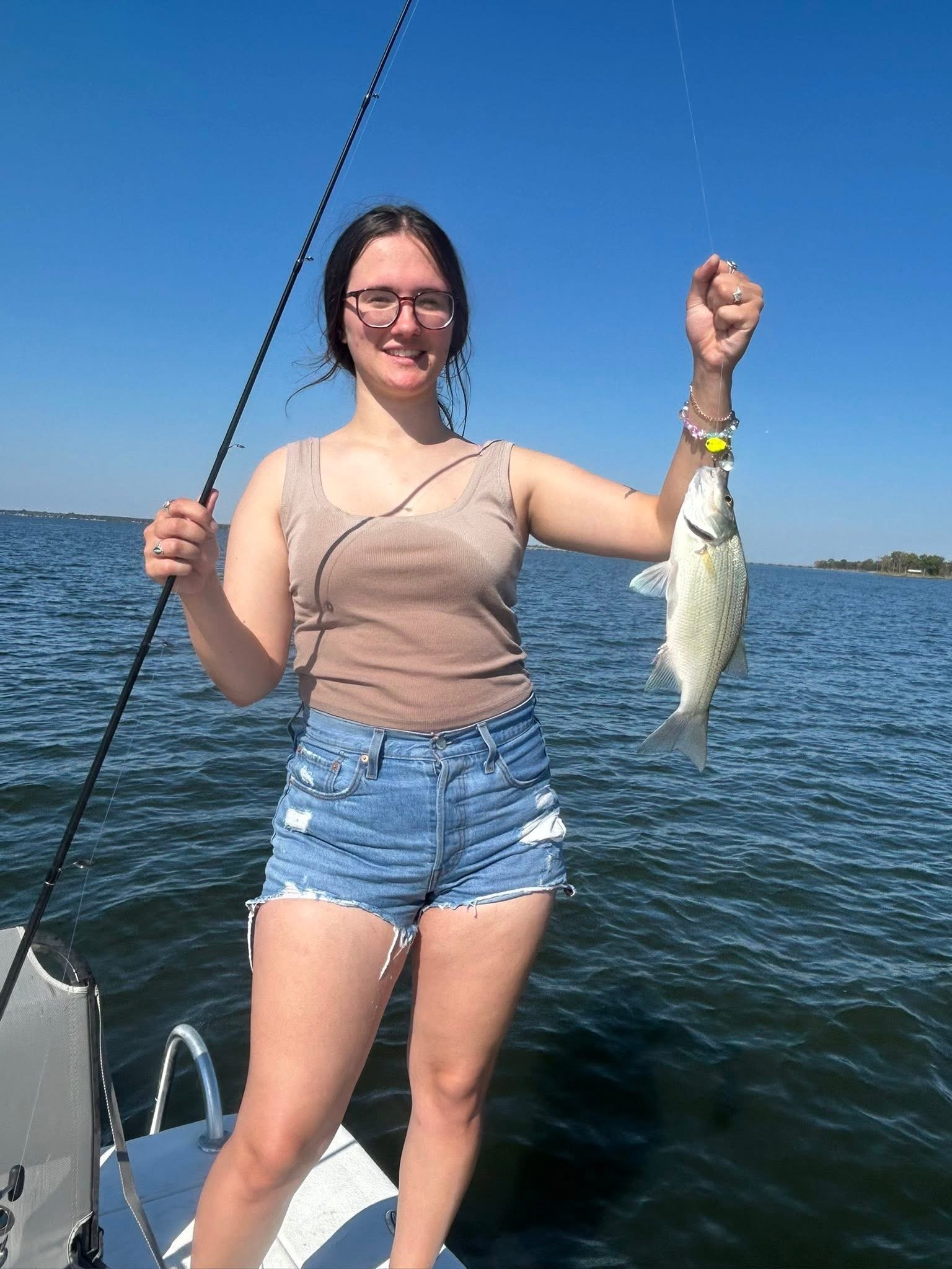 A young woman with glasses and dark hair holding a fish on a fishing boat in a body of water under a clear blue sky.
