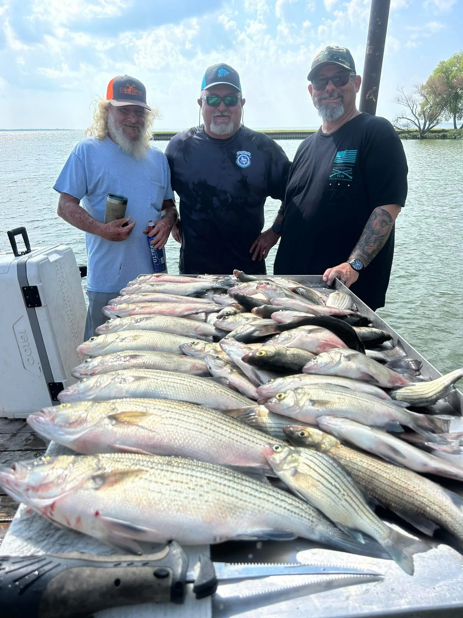 Three men standing on a boat with a large catch of fish spread on a table in front of them, with water and trees in the background.