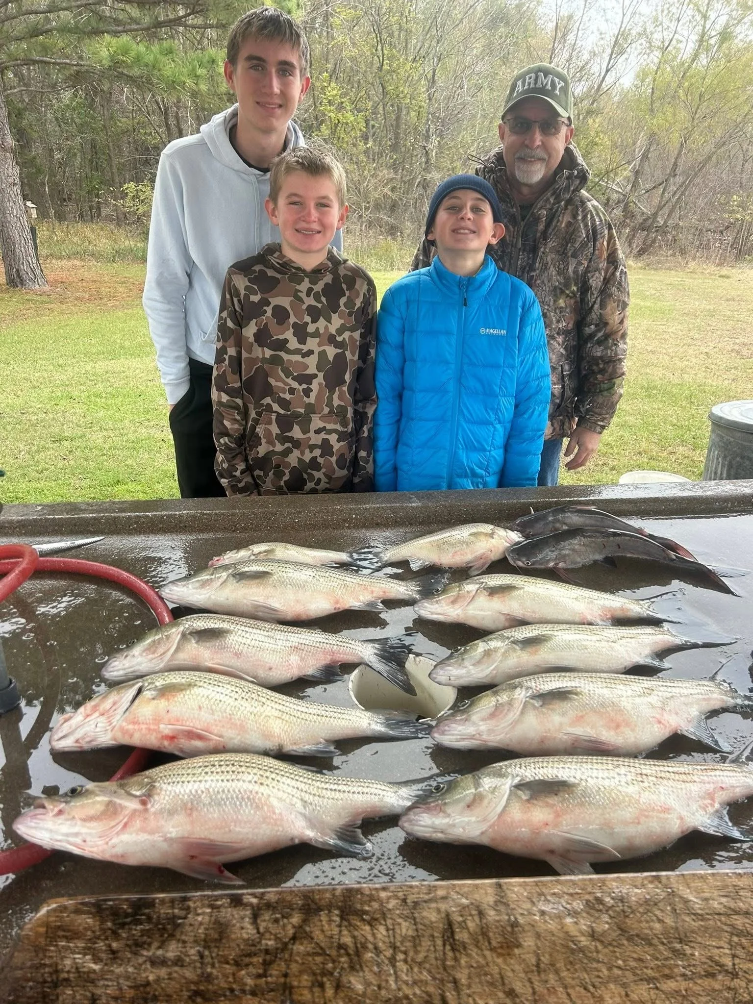 A group of four people standing outdoors behind a table with freshly caught fish, with trees and grass in the background.