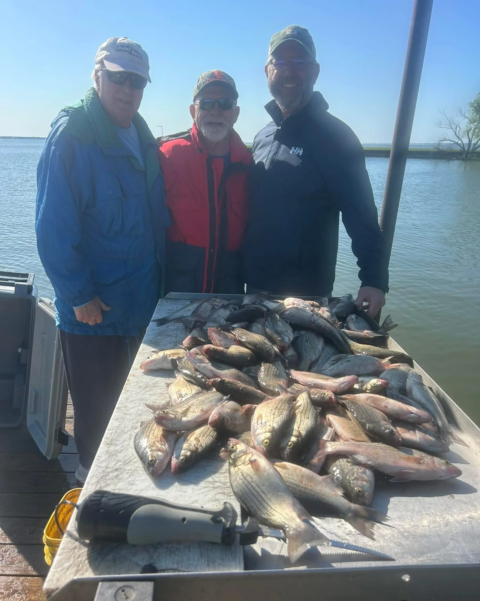Three men in outdoor clothing and hats standing behind a table full of fish, with a body of water and clear sky in the background, suggesting a fishing trip.