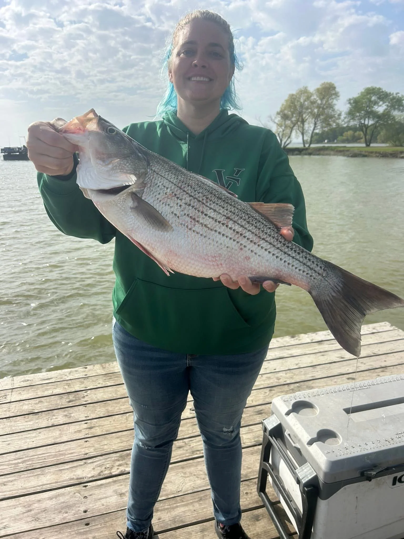 A woman with blue hair holding a large striped fish at a dock, with water and trees in the background.