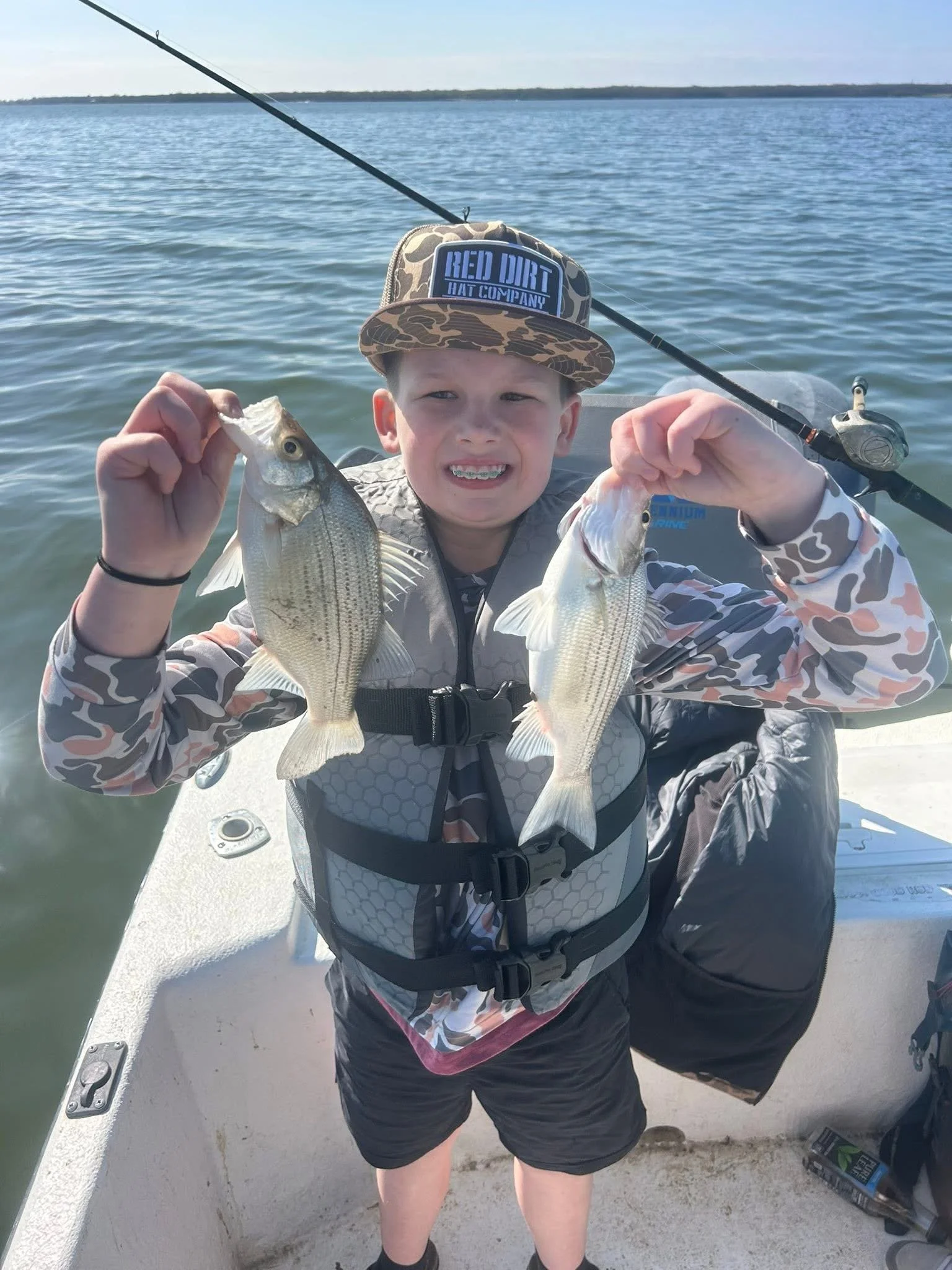 Young boy on a boat holding two fish, smiling, with fishing gear and water in the background.