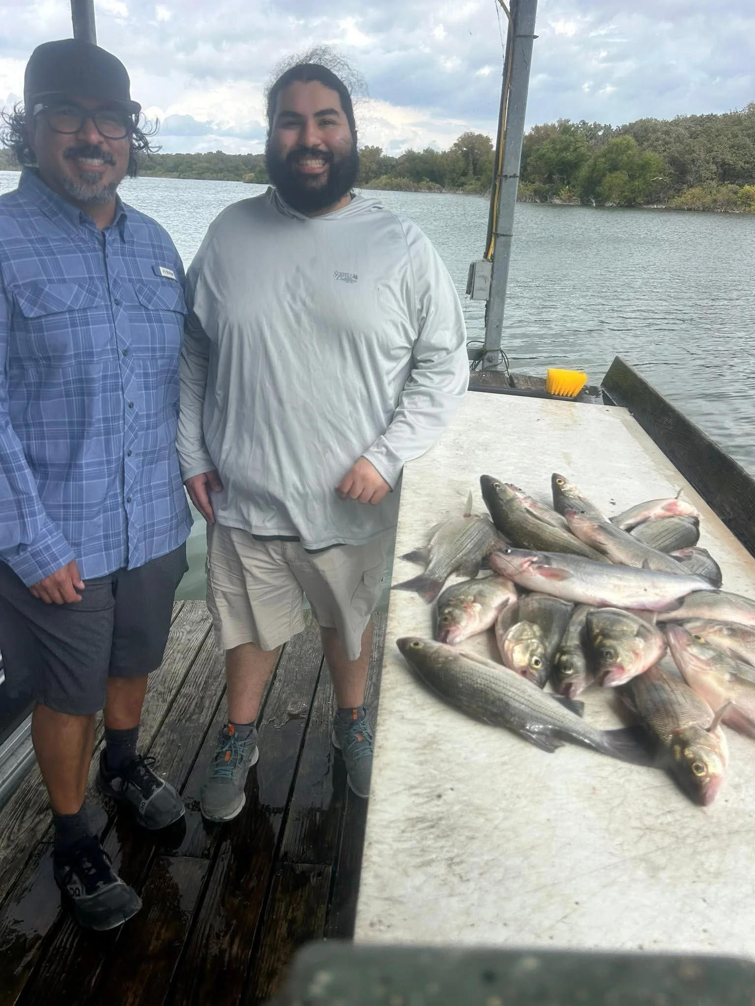 Two men standing on a dock by the water with a large catch of fish laid out on a table in front of them.
