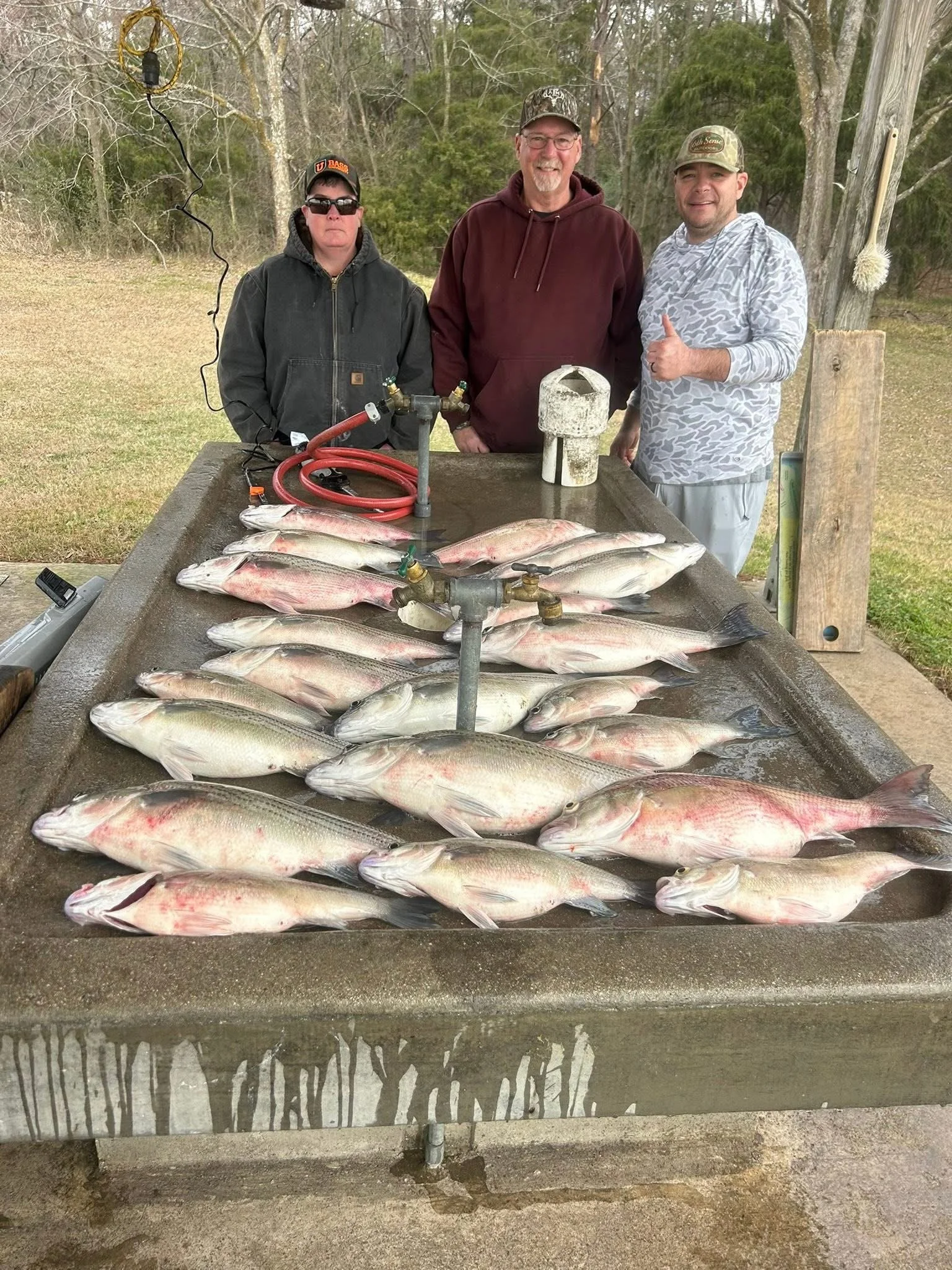 Three men standing outdoors behind a large catch of fish laid out on a table with multiple water spigots. The men are smiling, one giving a thumbs-up, and the background features trees and a wooden post with a brush hanging from it.