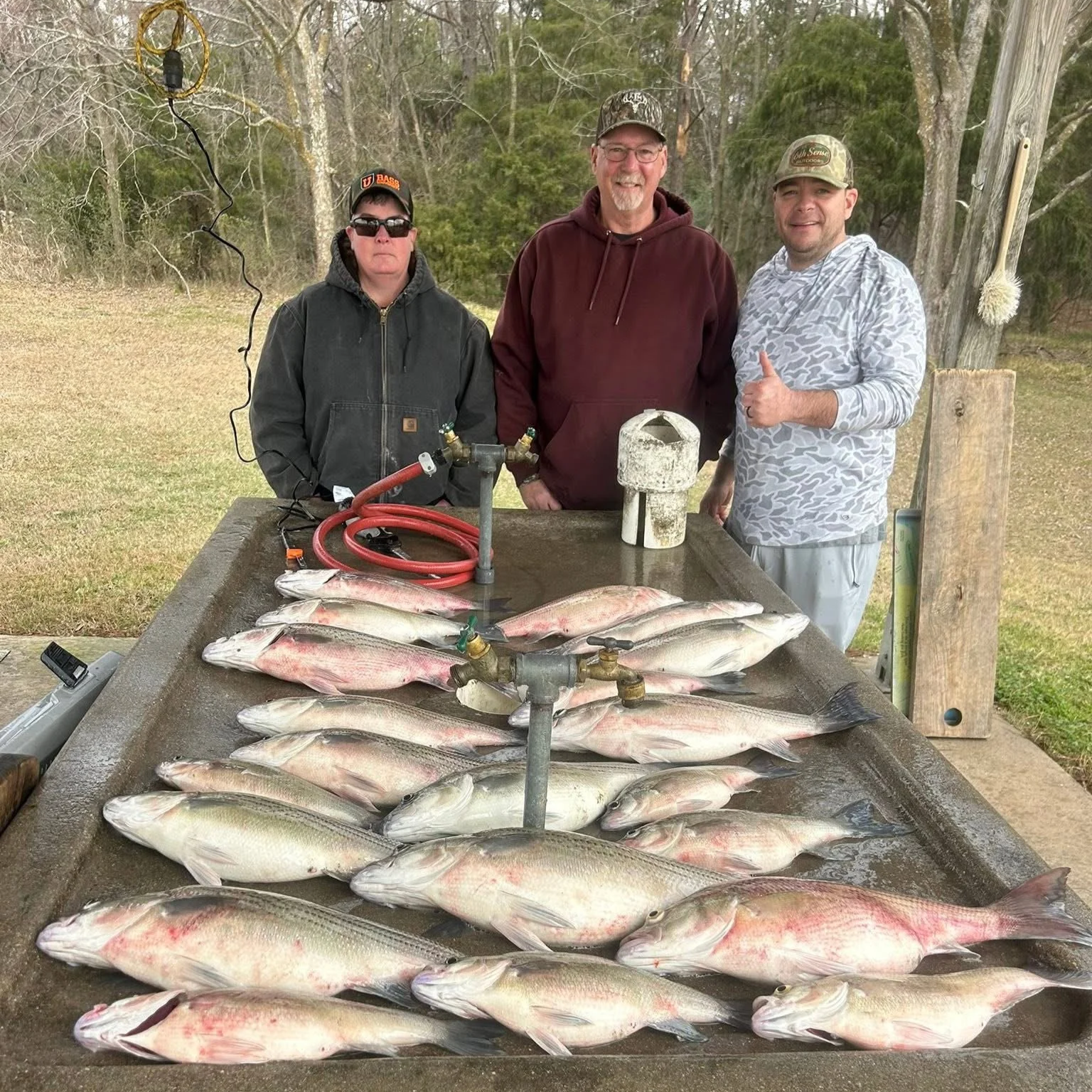 Three men standing behind a table with freshly caught fish laid out in rows. The table has a water faucet and some cleaning equipment. The men are outdoors, in a natural setting with trees in the background. Two of the men are smiling, one giving a t