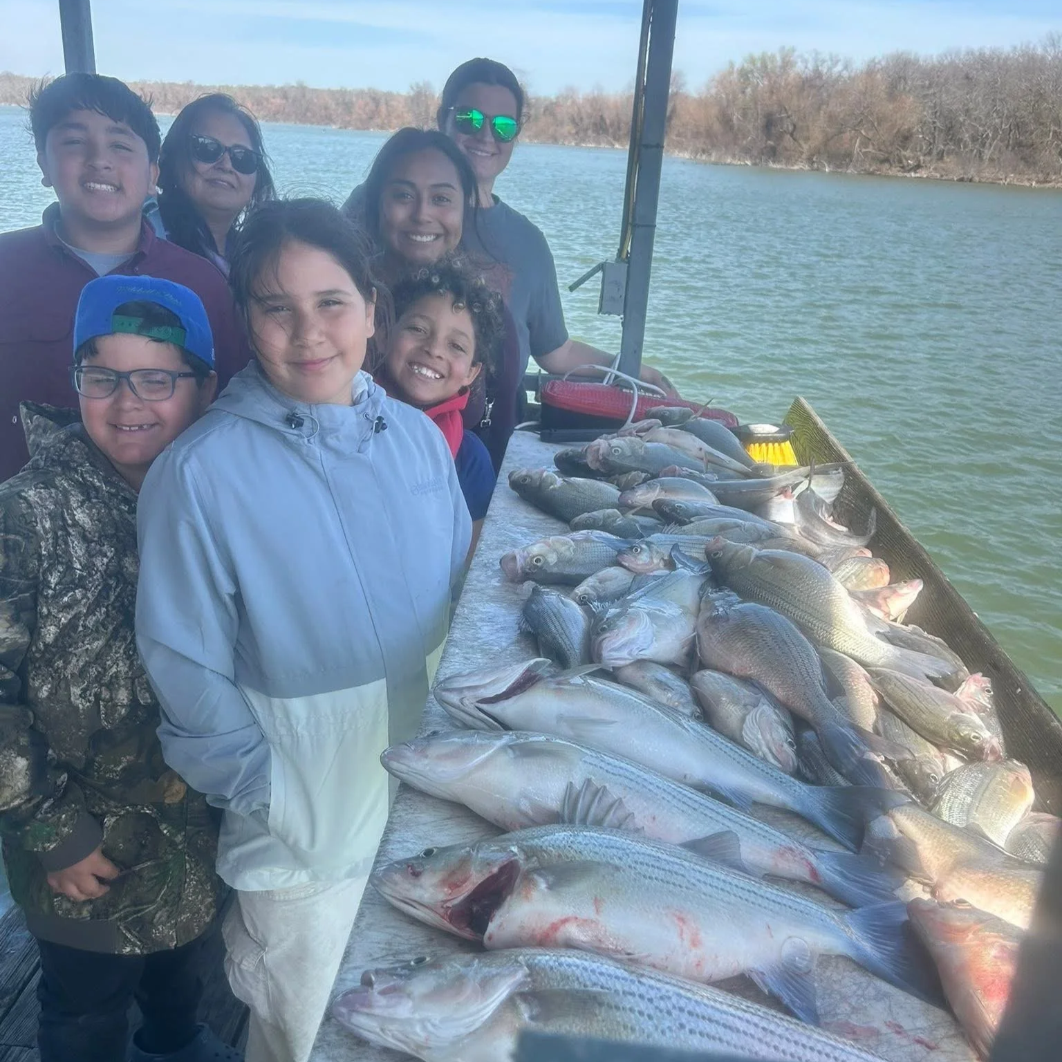 Children and adults on a boat by the water with a large catch of fish laid out on a table, smiling at the camera.