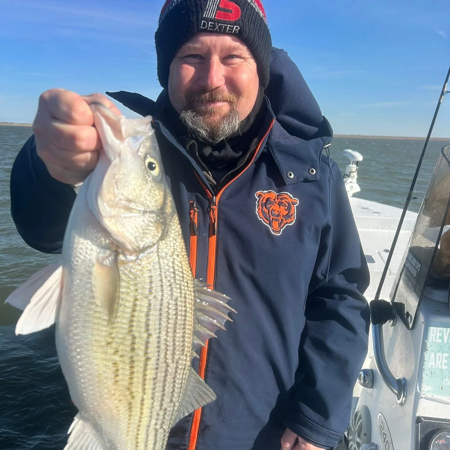 A man wearing a black beanie and a dark jacket with a tiger logo, holding a fish he caught on a boat in open water during daytime.