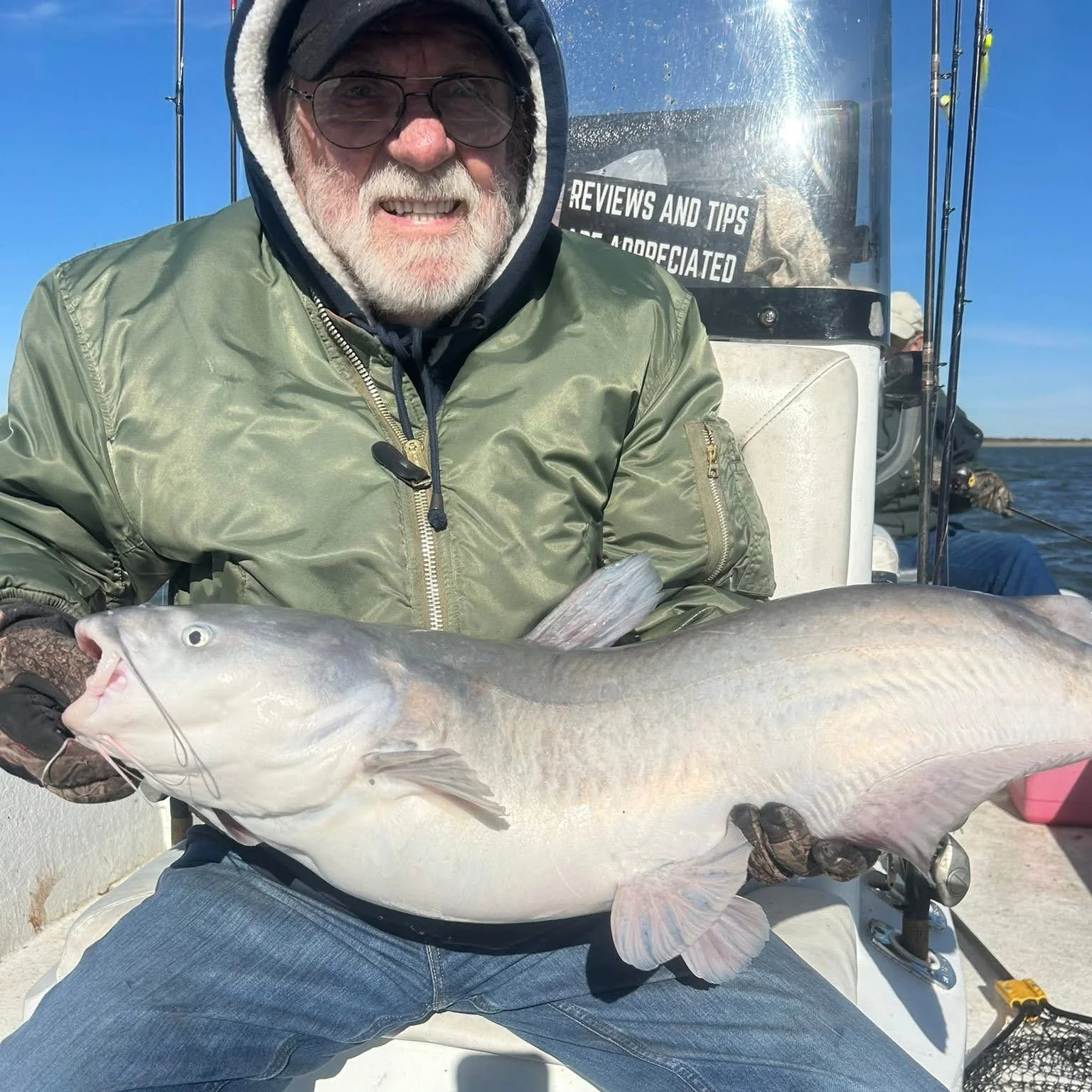 A man in a green jacket and glasses holding a large fish on a boat with water and blue sky in the background.