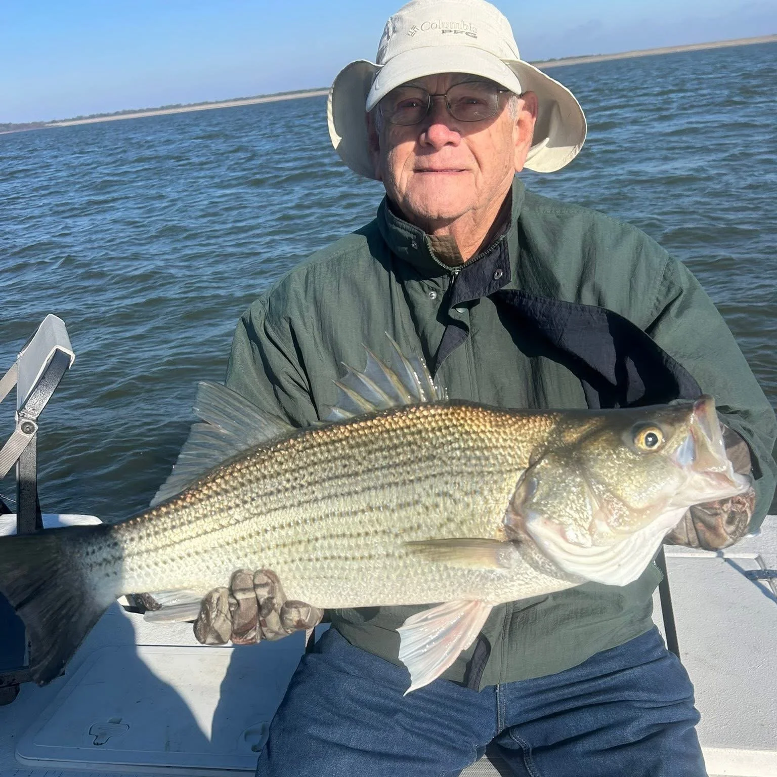 An elderly man wearing a beige hat and glasses holds a large fish on a boat in the water with a distant shoreline in the background.