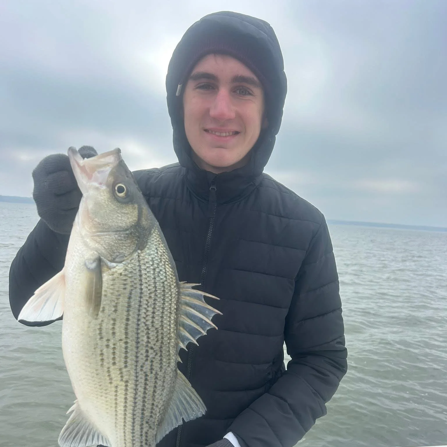 A young man in a black jacket and black beanie holding a large fish, standing on a body of water with an overcast sky in the background.