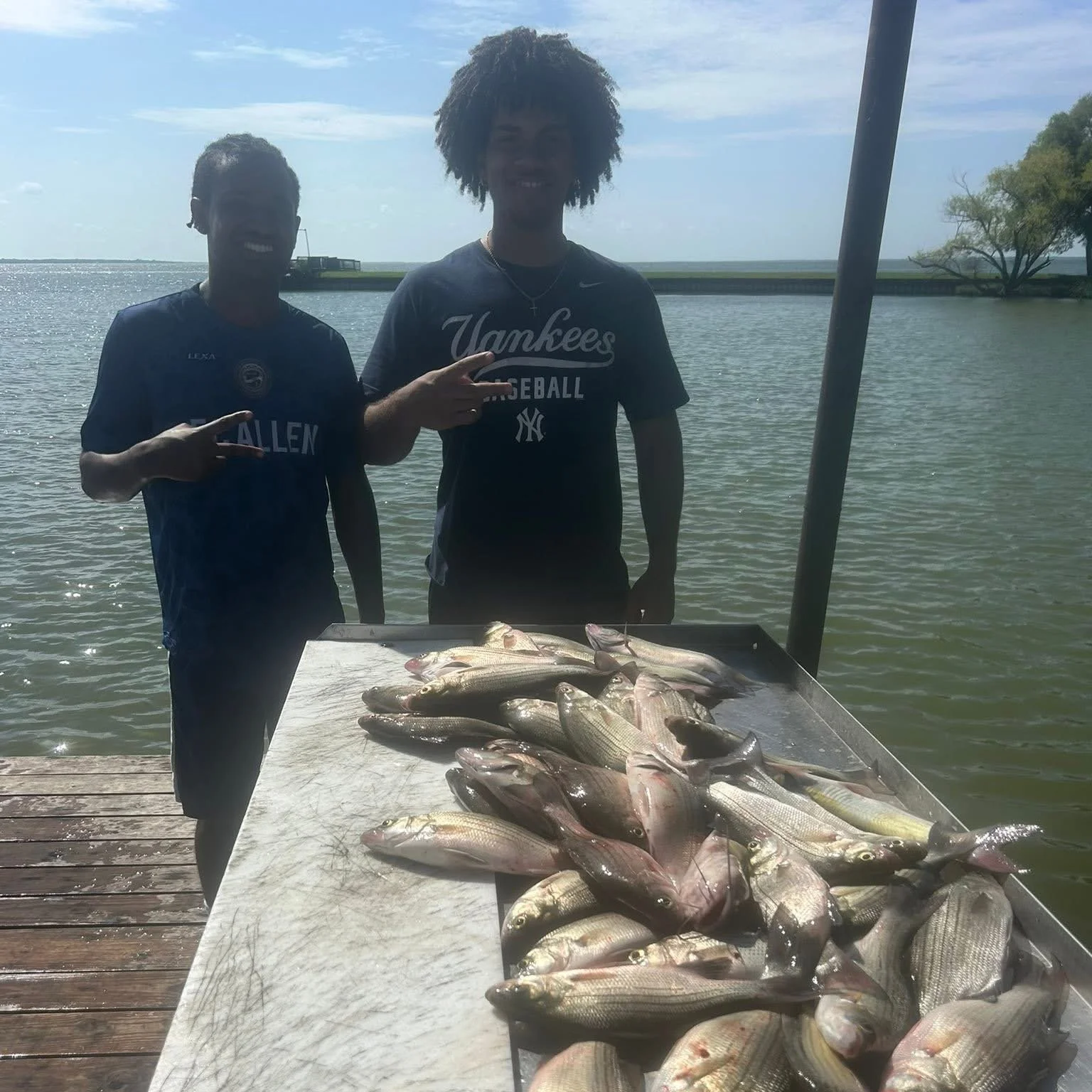 Two young men standing behind a table with freshly caught fish, outdoors near the water with a dock and trees in the background.