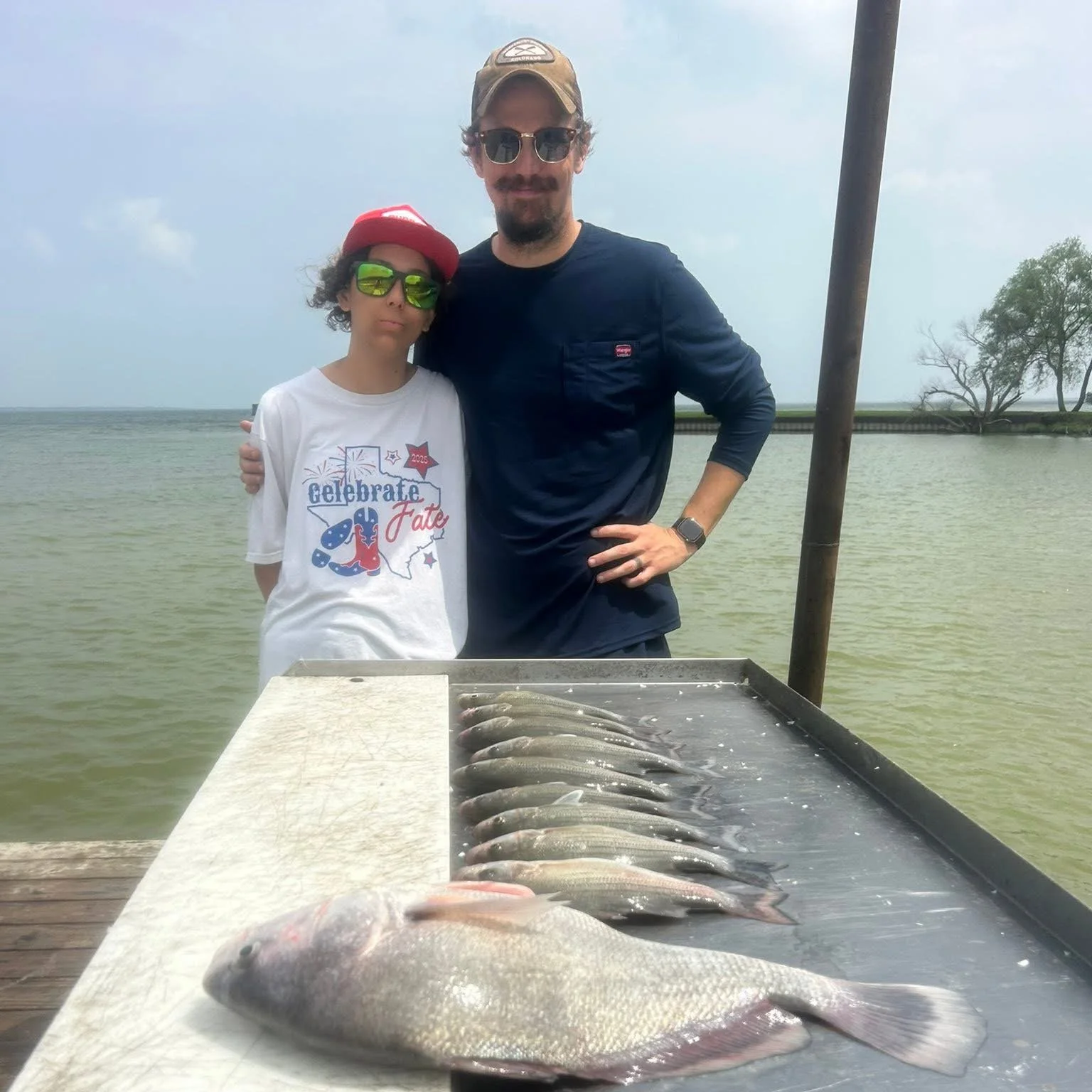 Two people standing behind a table with fish on it at a waterfront, with water, a tree, and a cloudy sky in the background.