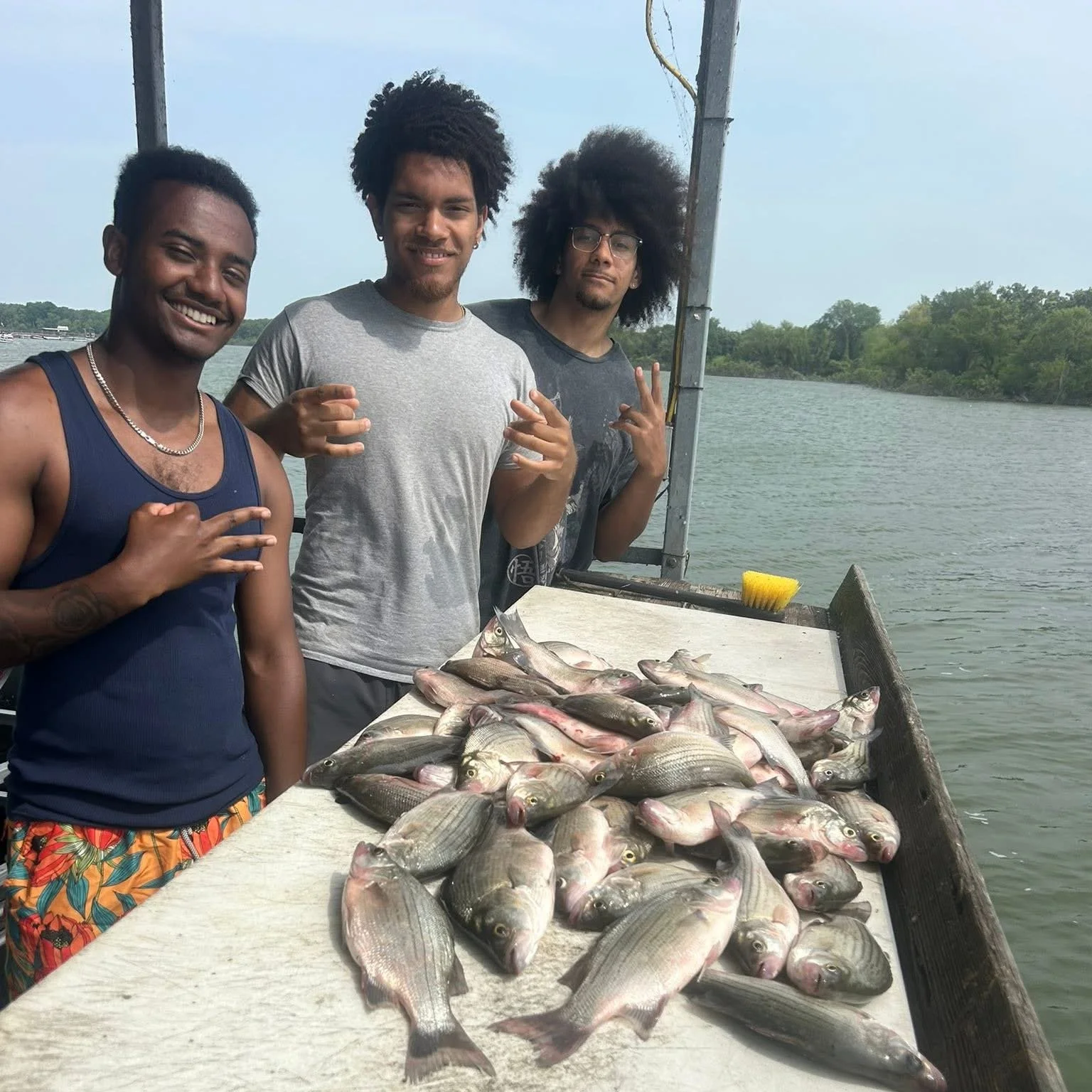 Three men standing behind a table of fish by a body of water, with a fishing boat in the background, suggesting a fishing trip or catch.