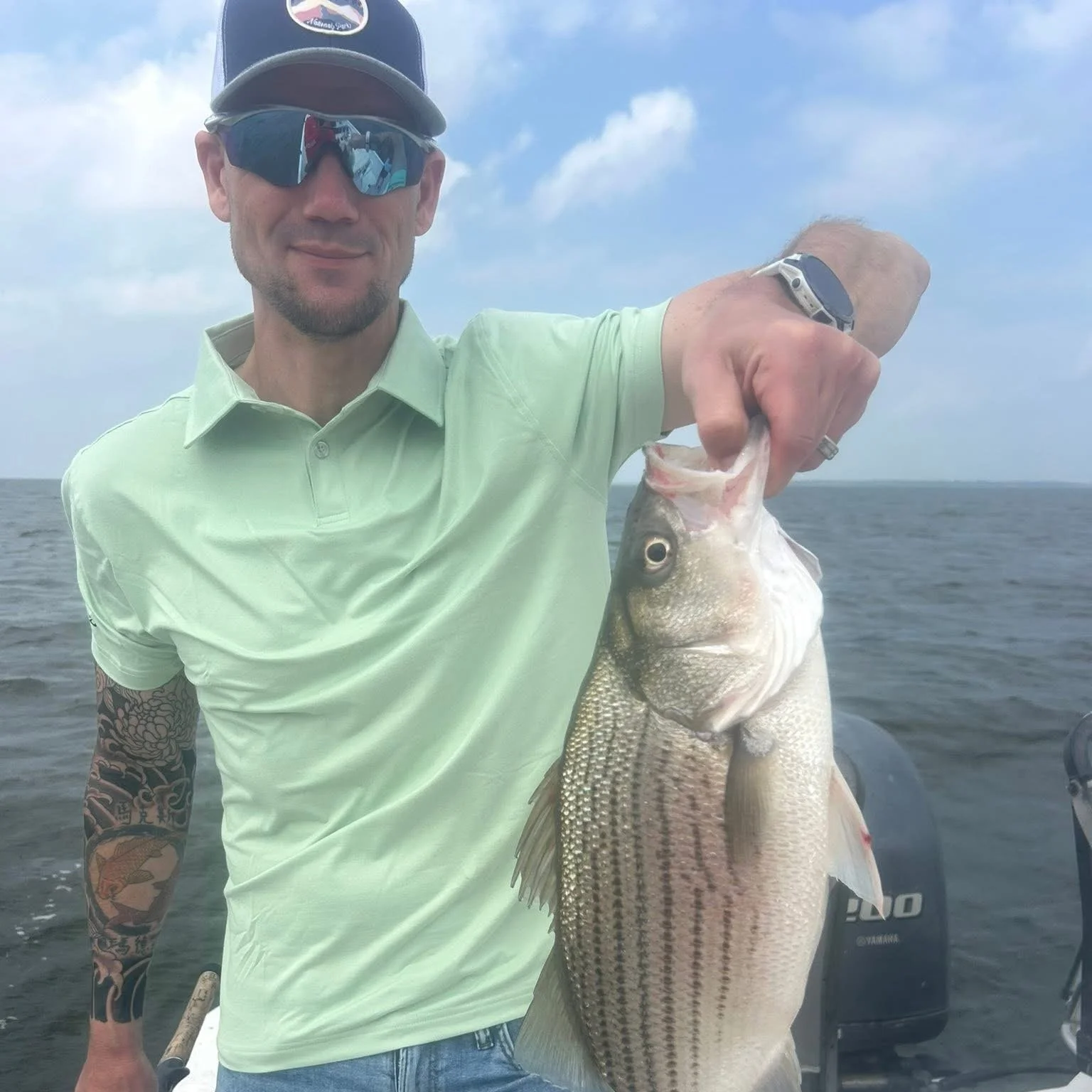 Man holding a large fish on a boat, wearing sunglasses, a cap, and a tattoos on his arm, with water and sky in the background.
