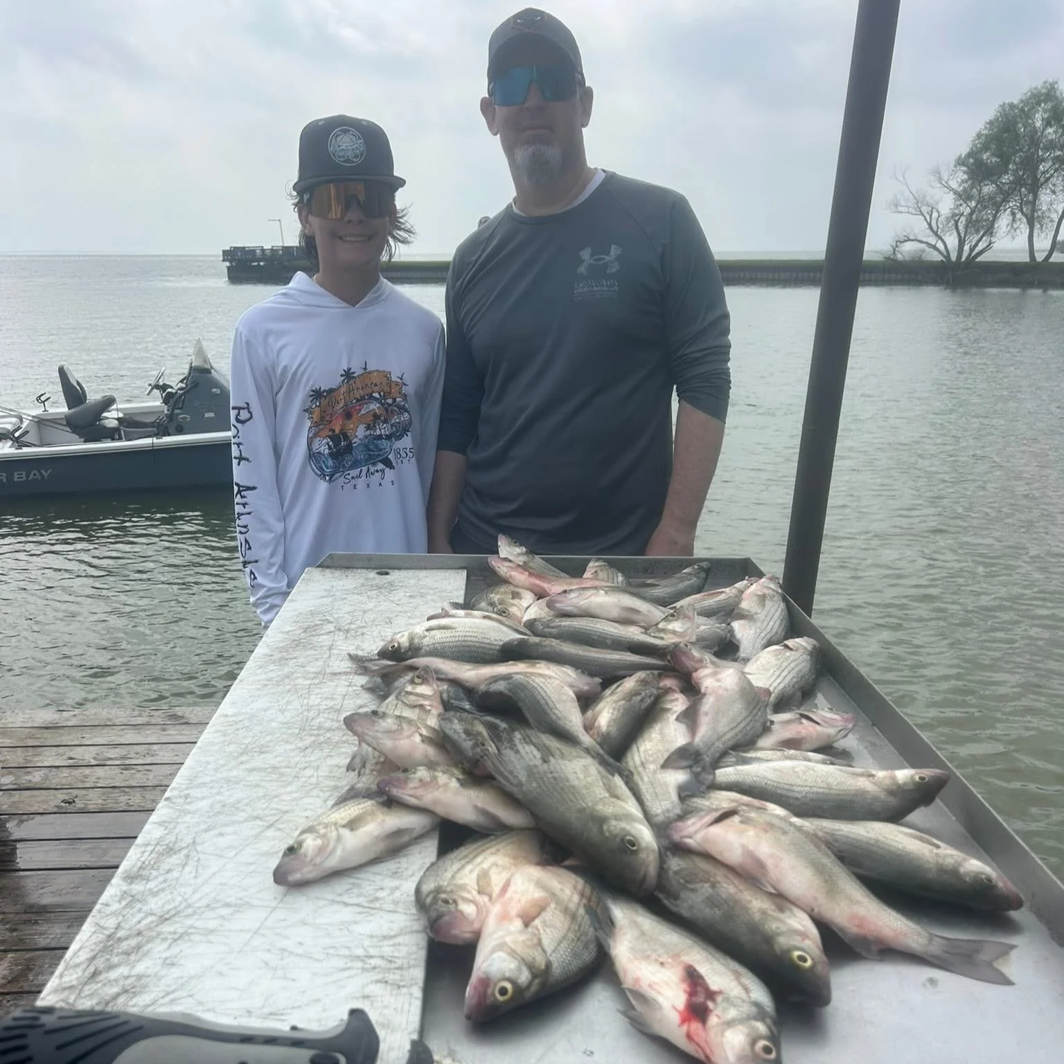 Two men standing by a table full of freshly caught fish, with a boat and water in the background, on a cloudy day.