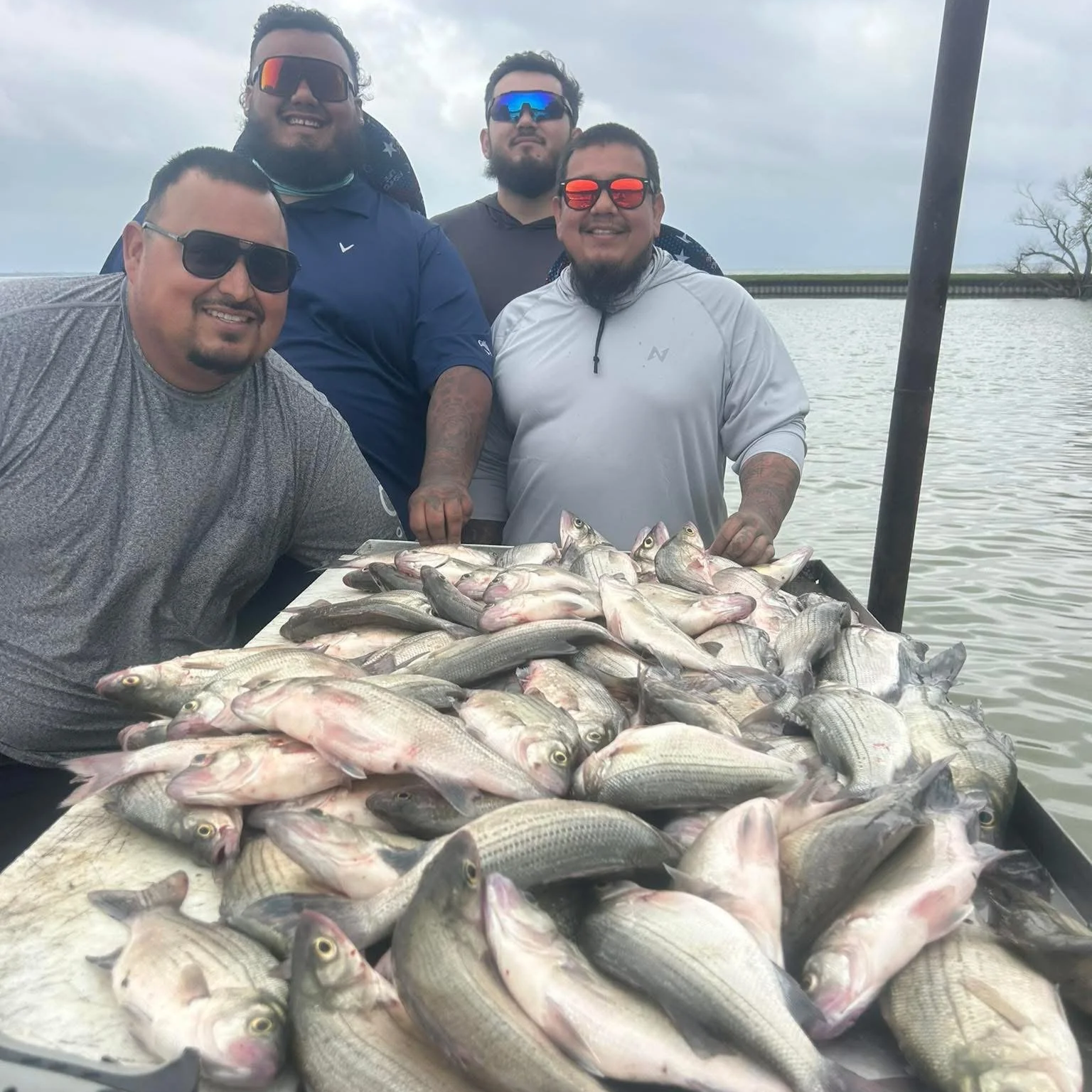 Group of five men smiling, displaying a large catch of fish on a table near a body of water under a cloudy sky.