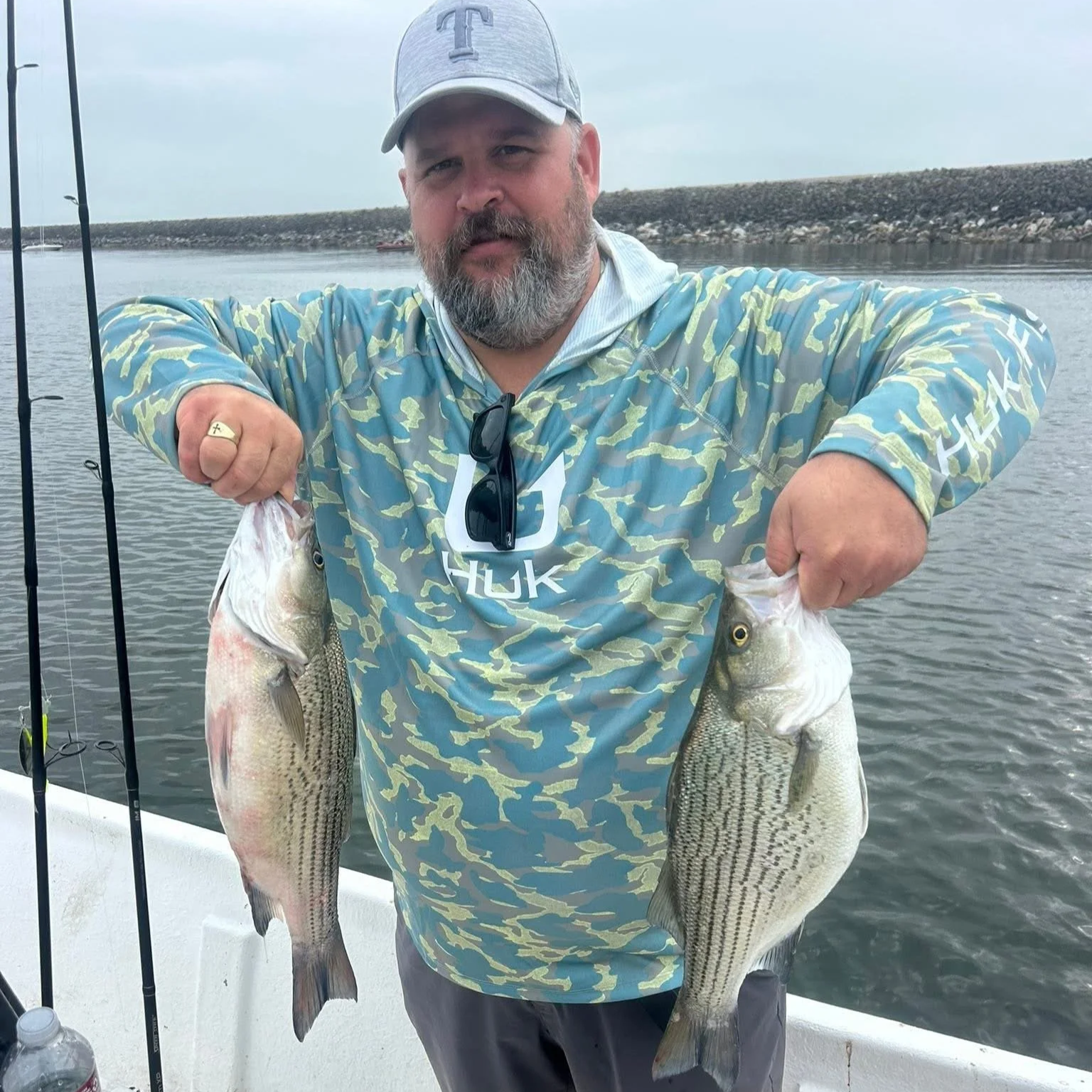 A man with a beard wearing a gray cap and a camouflage hoodie holding two large fish, one in each hand, standing on a boat docked in a body of water with rocks and a breakwater in the background.