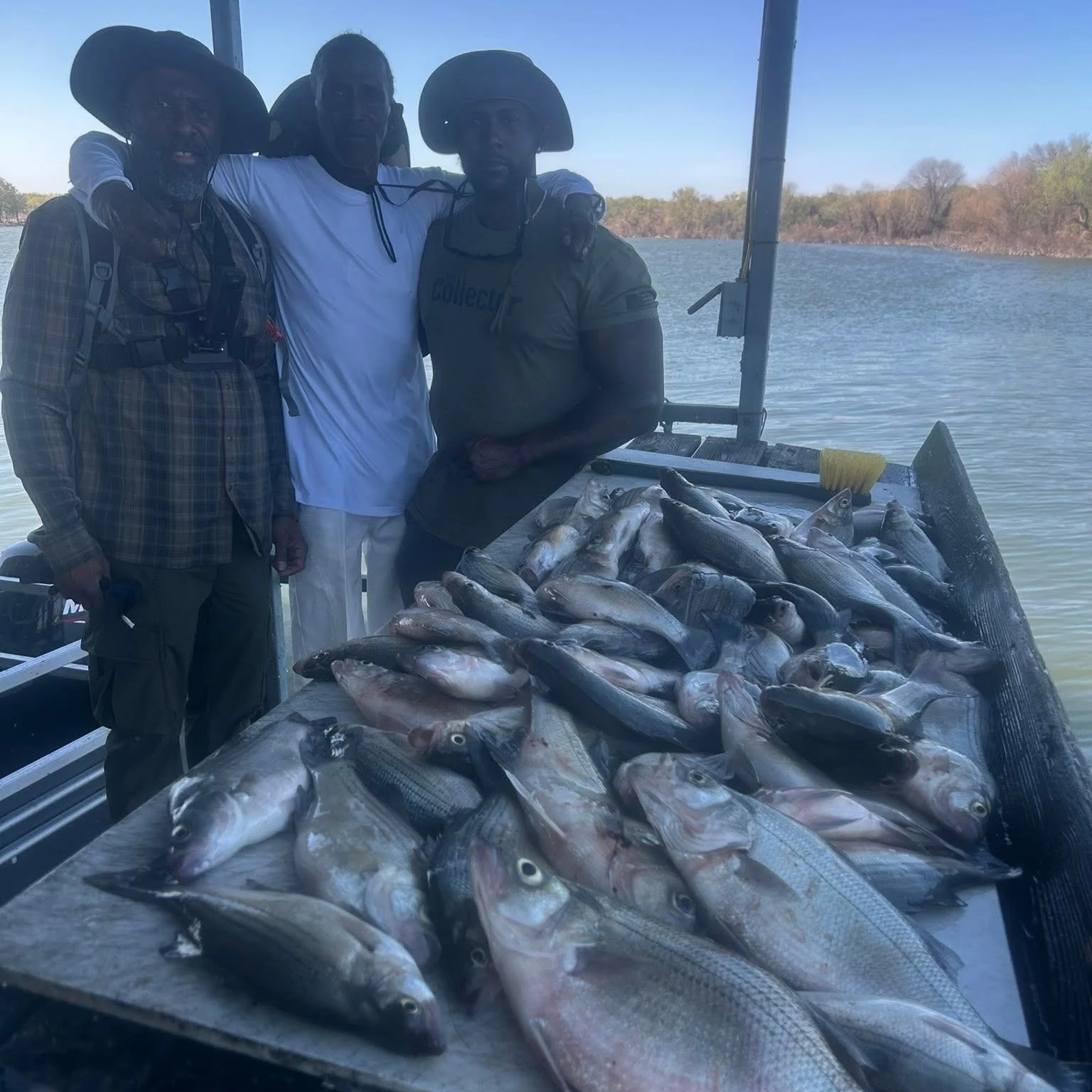 Three men standing on a boat with a large catch of fish laid out on a table. The men are outdoors near a body of water with trees in the background.