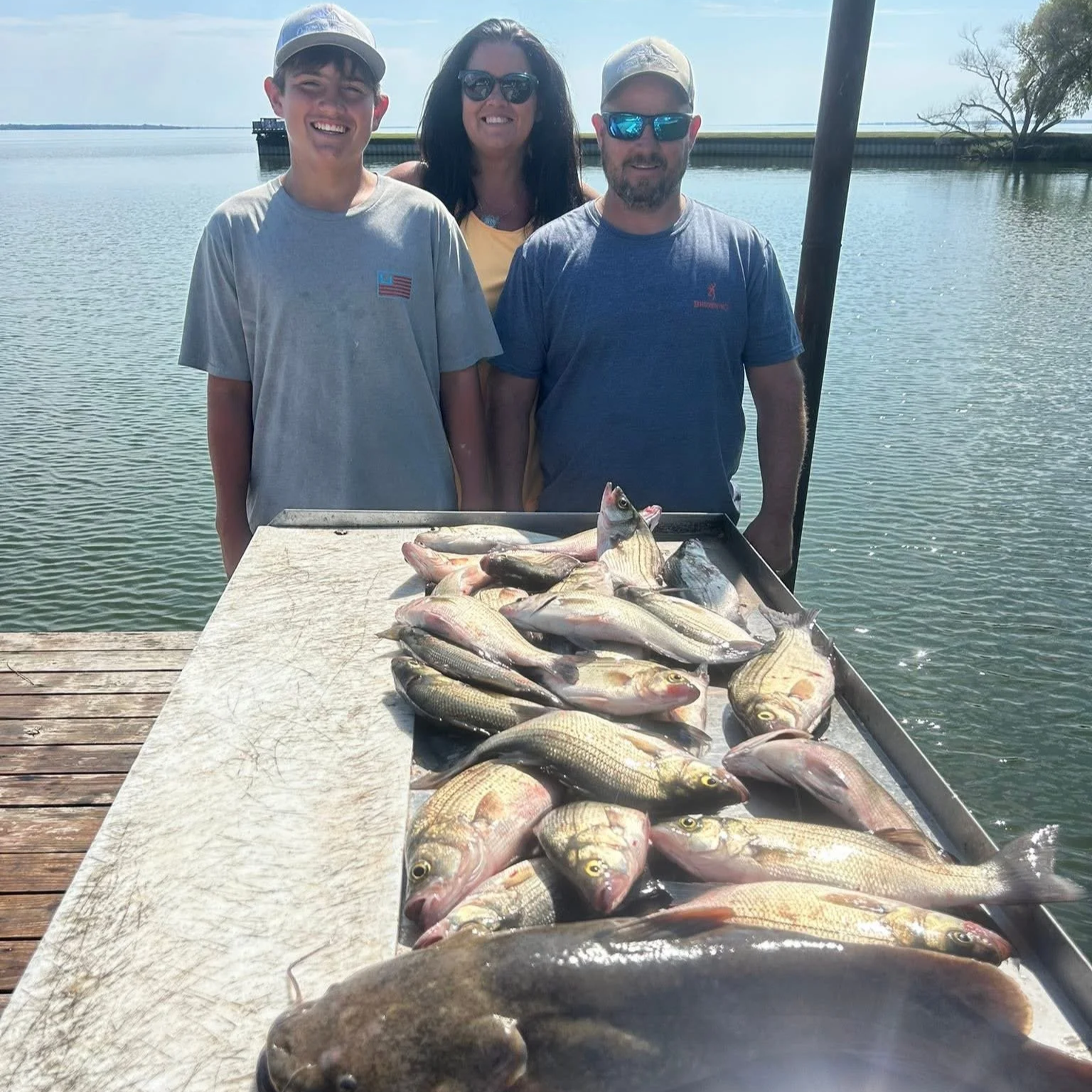 Three people standing behind a table filled with a large catch of fish, on a dock near a body of water with a dock and some trees in the background.