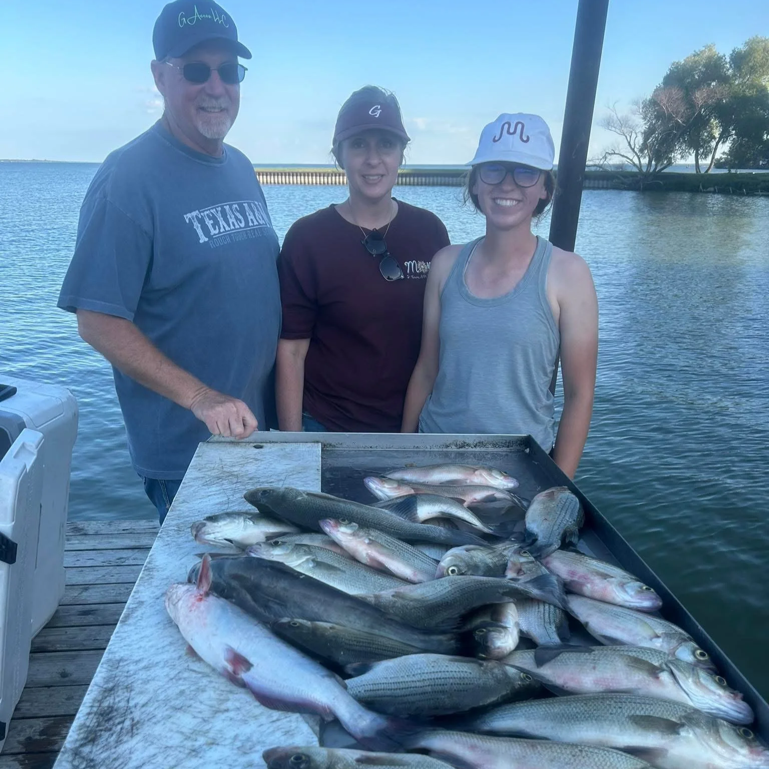 Three people standing behind a table full of fish on a dock by the water, with a backdrop of a river and trees, enjoying a fishing trip.