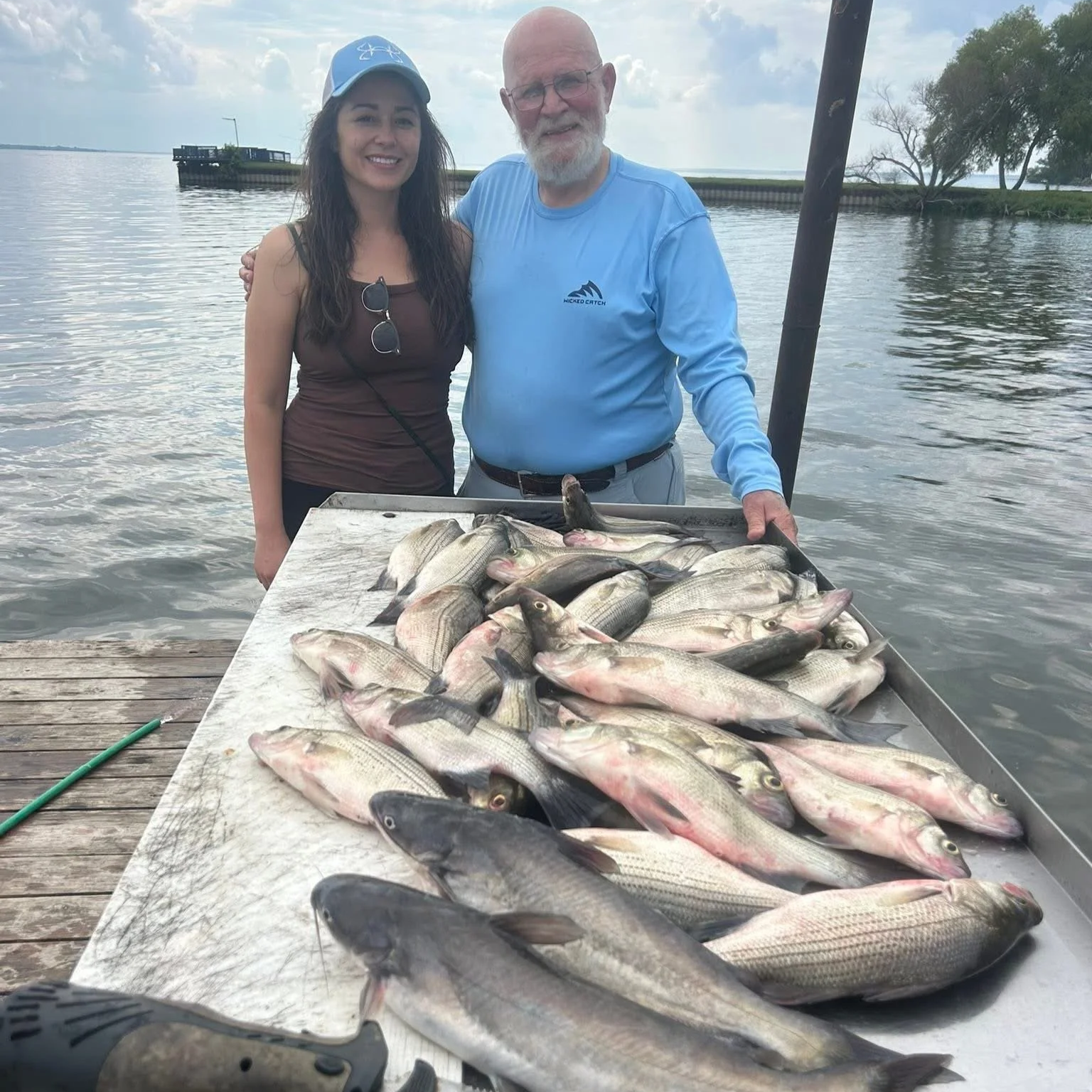 A young woman and an older man with a beard and glasses stand by a table of many fish caught from a lake or river, with water and trees visible in the background.