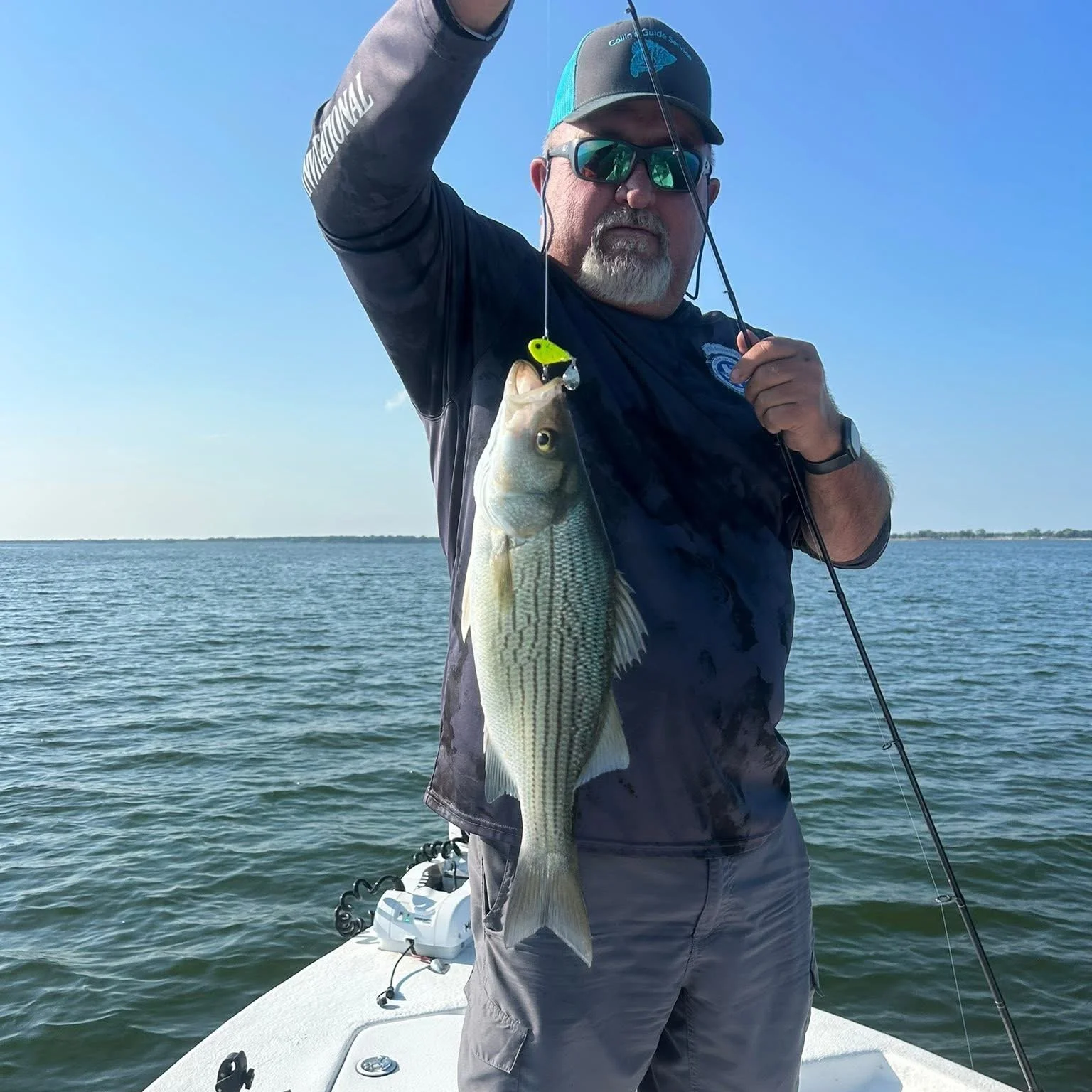 A man in sunglasses and a cap holding a large fish on a fishing line on a boat on a body of water with a clear sky.