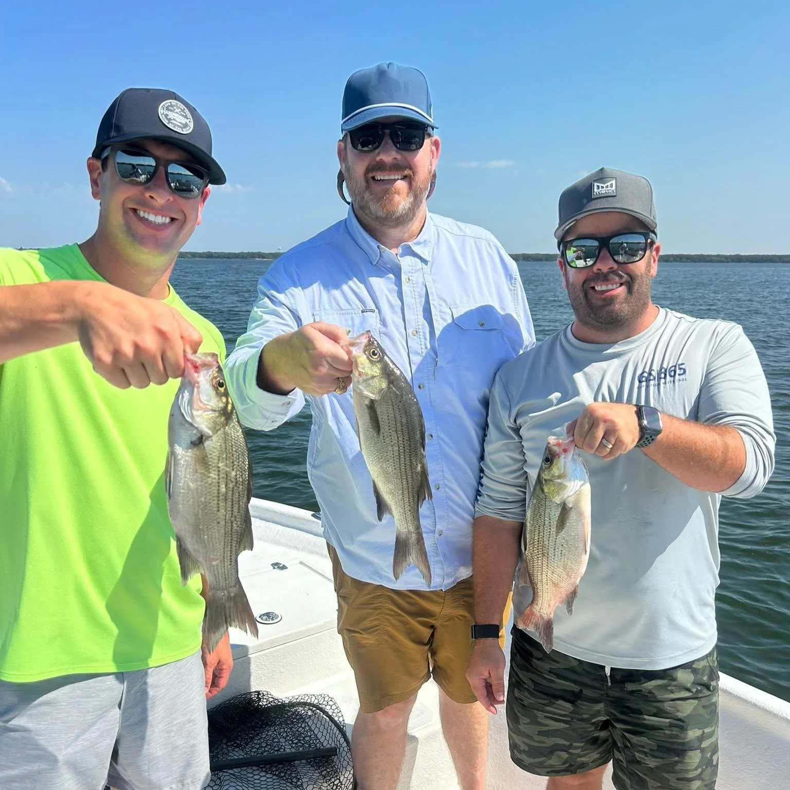 Three men on a boat holding fish they caught, with water and a clear blue sky in the background.