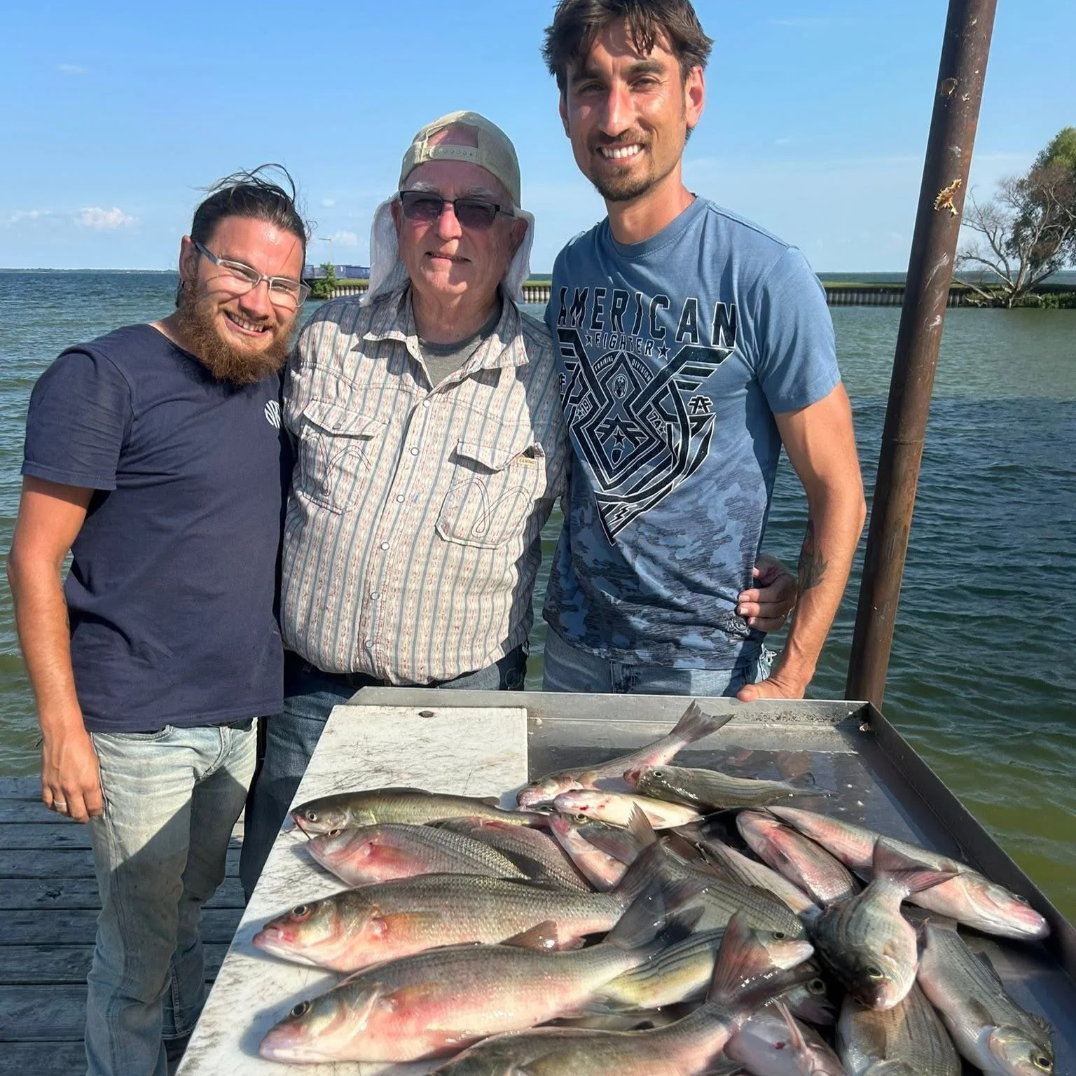 Three men standing on a dock with a table full of freshly caught fish, with water and sky in the background.