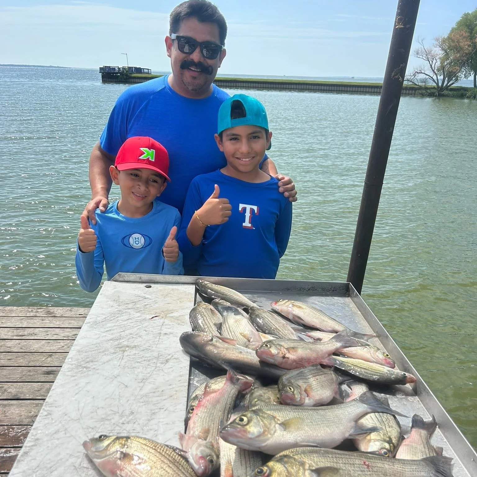 Three people, a man and two boys, stand behind a table filled with caught fish near a body of water. They are outdoors, on a dock or pier, and appear happy, with the boys giving thumbs-up. The man is wearing sunglasses and a blue shirt, and the boys 