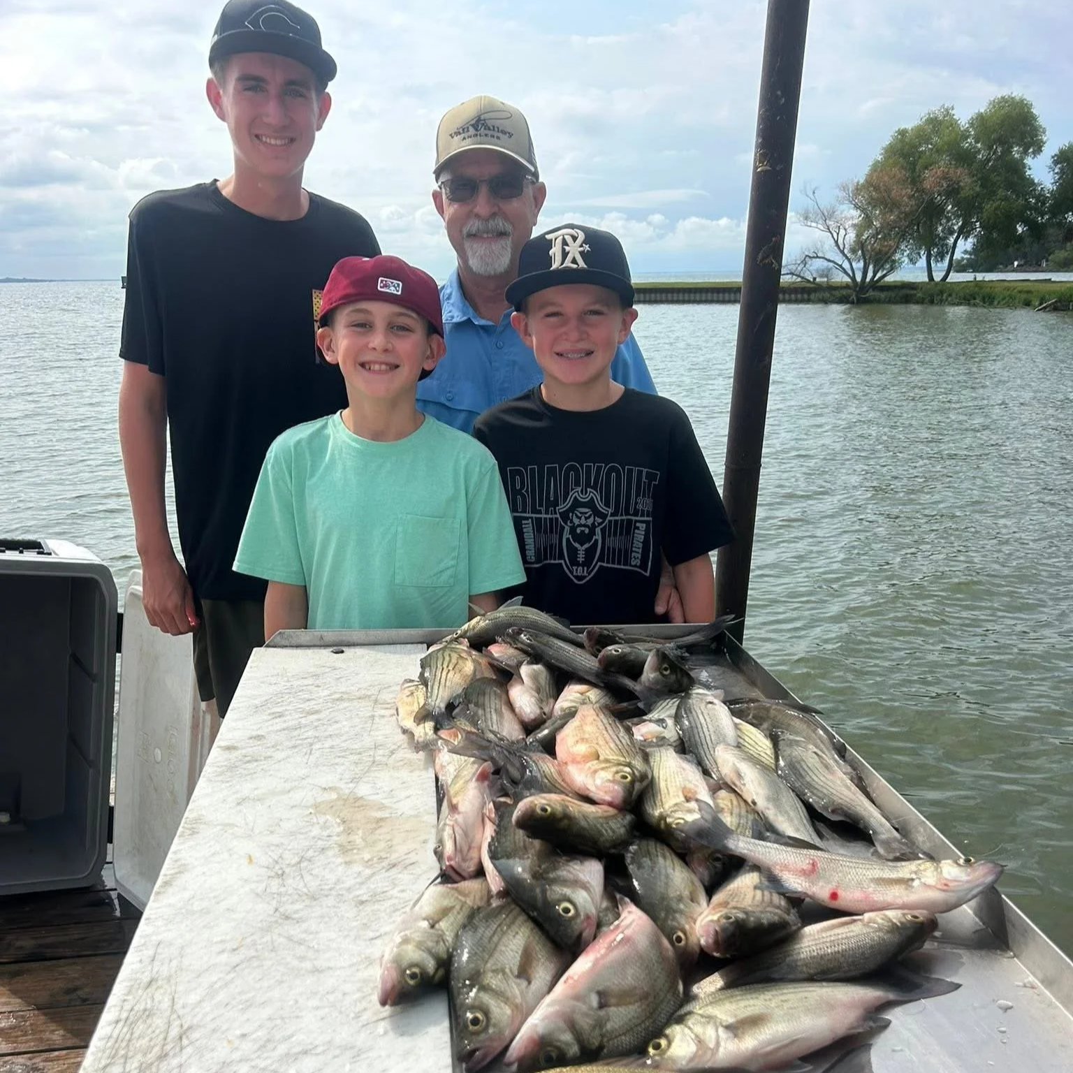 A group of four people, two children and two adults, standing behind a table full of freshly caught fish, with a water body and cloudy sky in the background.