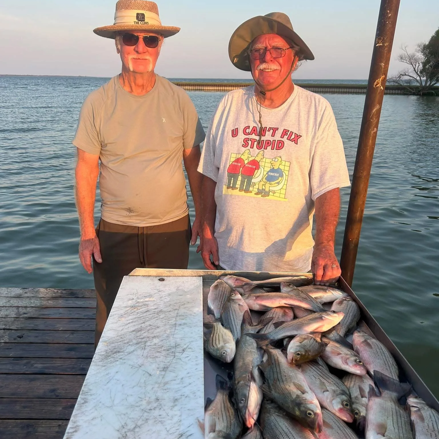 Two elderly men stand on a wooden dock near a body of water. One man wears a wide-brimmed hat, sunglasses, and a gray shirt, while the other wears a bucket hat and a T-shirt that says, 'U can't fix stupid' with cartoon characters. They are smiling, a