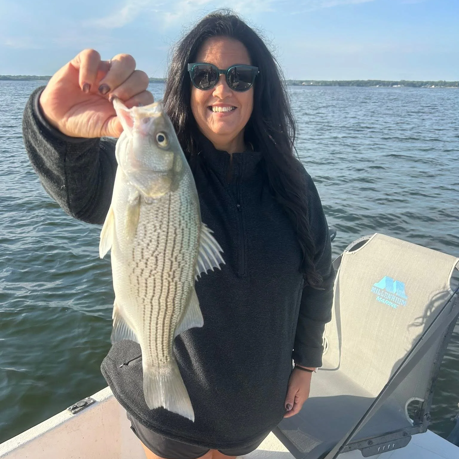 Woman on a boat holding a fish she caught, with water and a cloudy sky in the background.