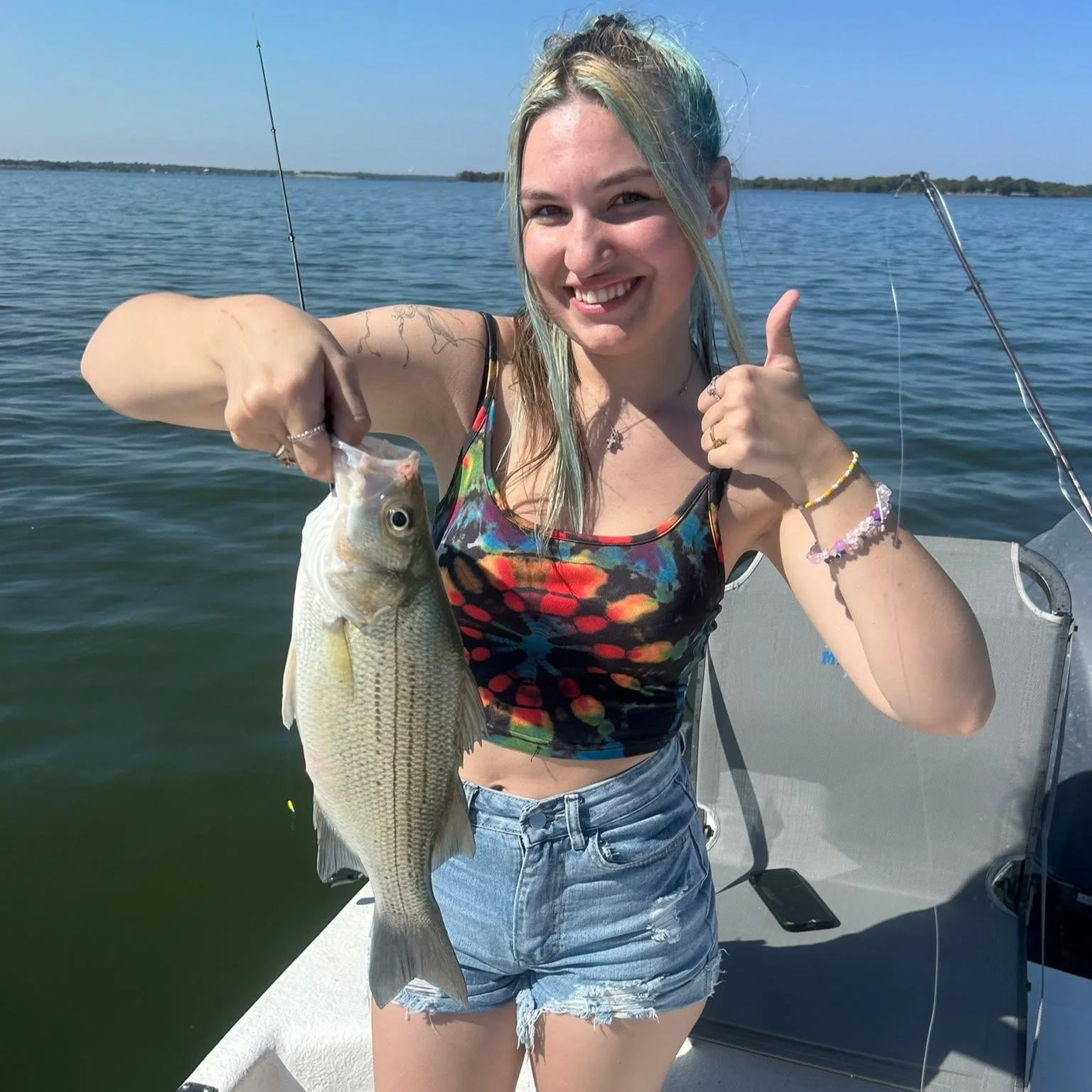 A young woman with multicolored hair, wearing a vibrant tank top and denim shorts, smiling and showing a thumbs up while holding a large fish she caught during a day of fishing on a boat in a calm lake or ocean.