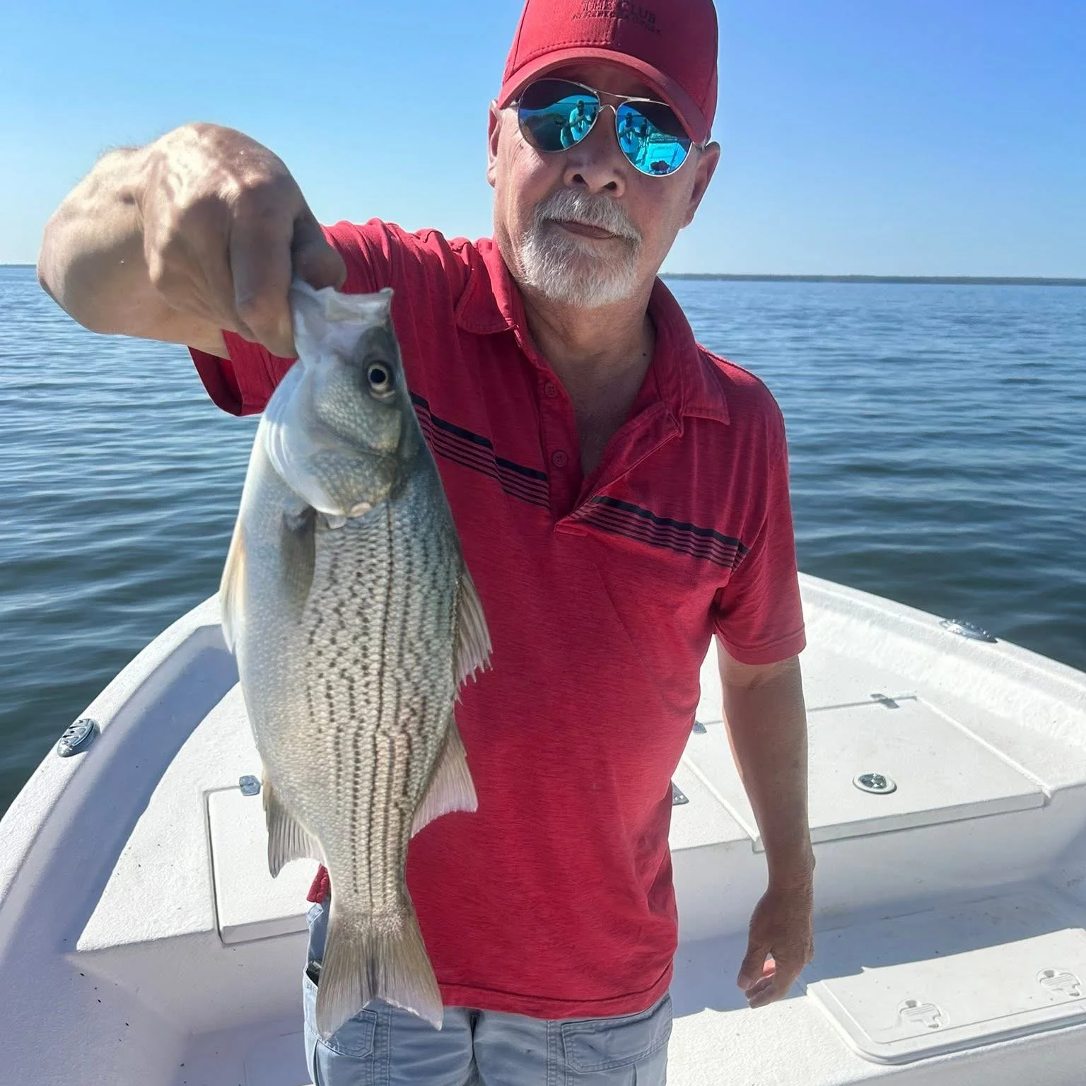 A man with a white beard and mustache wearing a red cap and sunglasses, holding a fish by its mouth on a boat in a body of water.