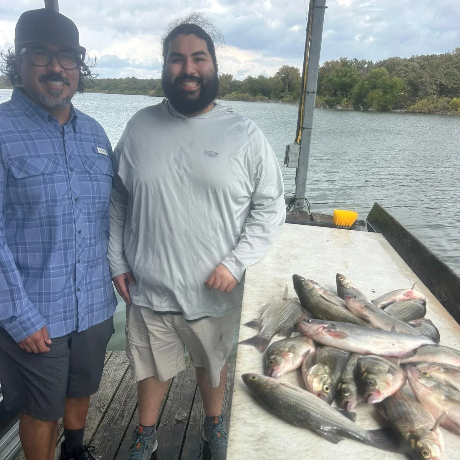 Two men smiling on a dock by a river with a table of freshly caught fish and a body of water with trees in the background.