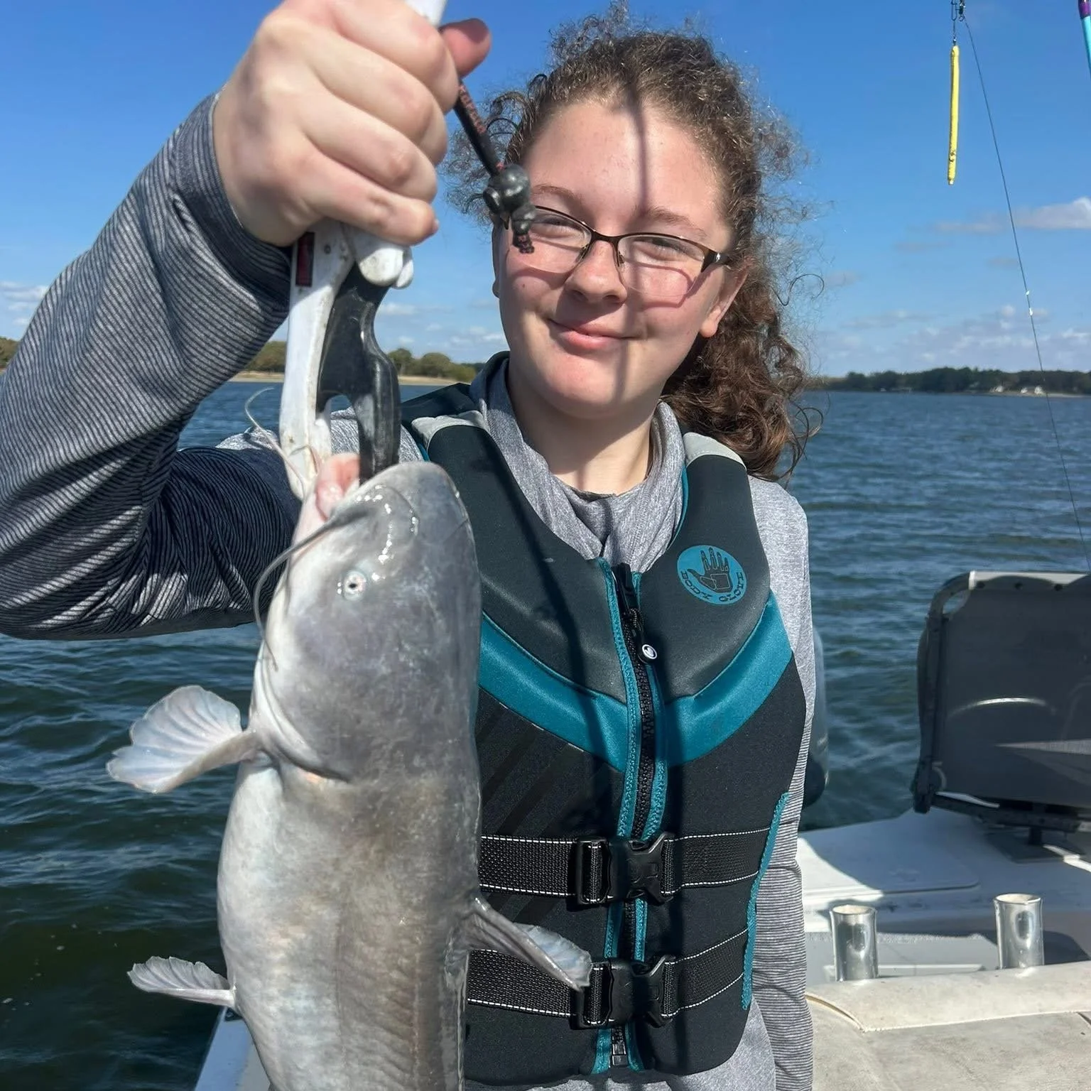 A young woman on a boat holding up a fish she caught, smiling at the camera. She is wearing glasses and a life jacket, with a water body and a blue sky in the background.