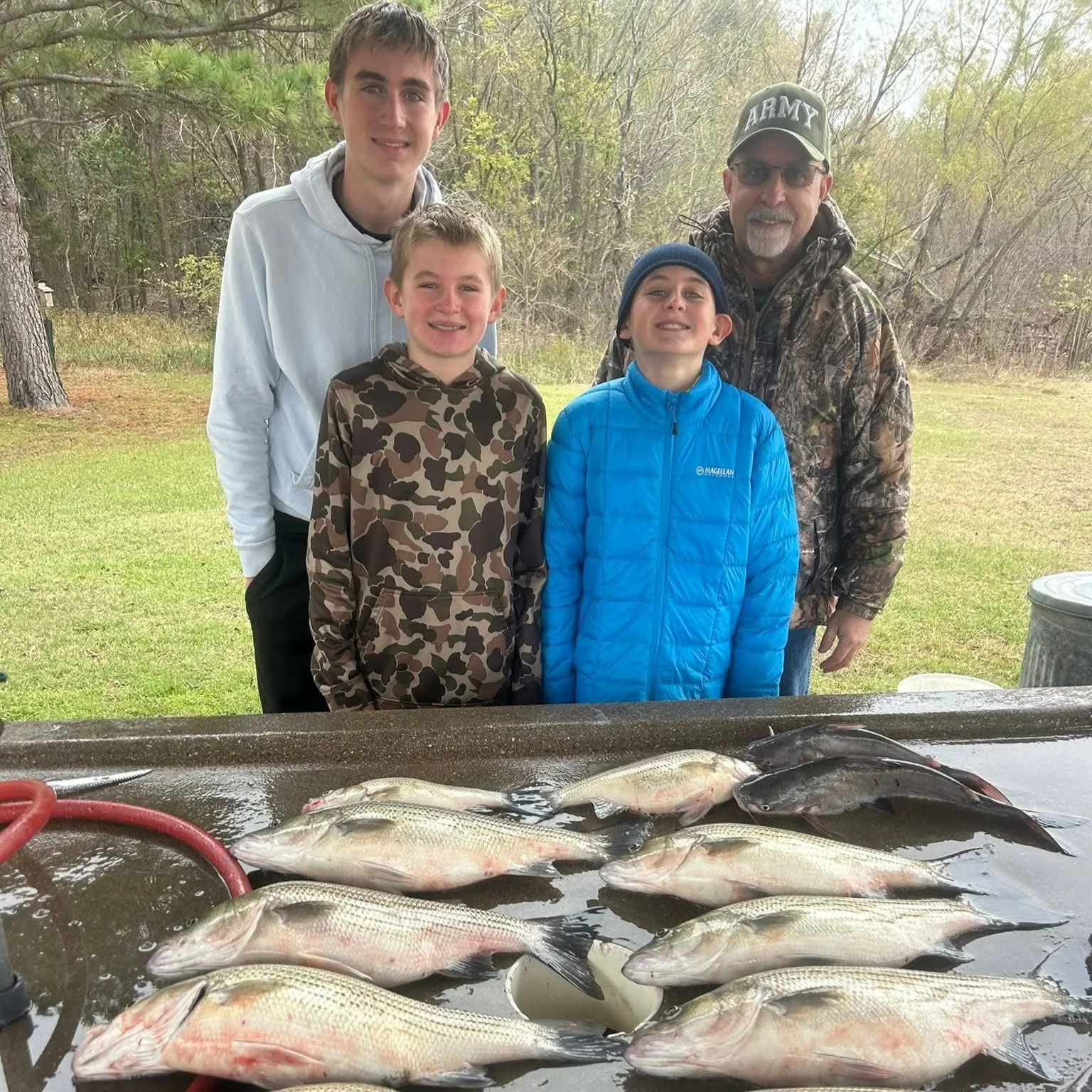 Four males standing outdoors behind a table with a large catch of fish, with trees and a grassy area in the background.