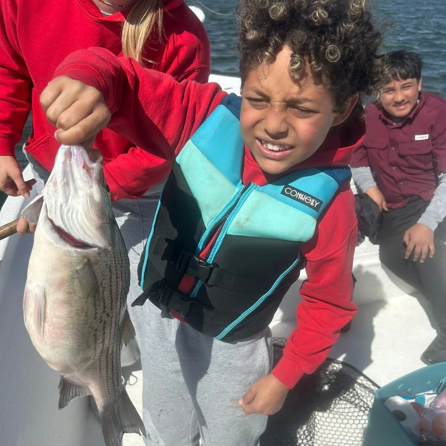 A young boy holding a large fish he caught, wearing a red jacket and a life jacket, with two other children in the background near water.